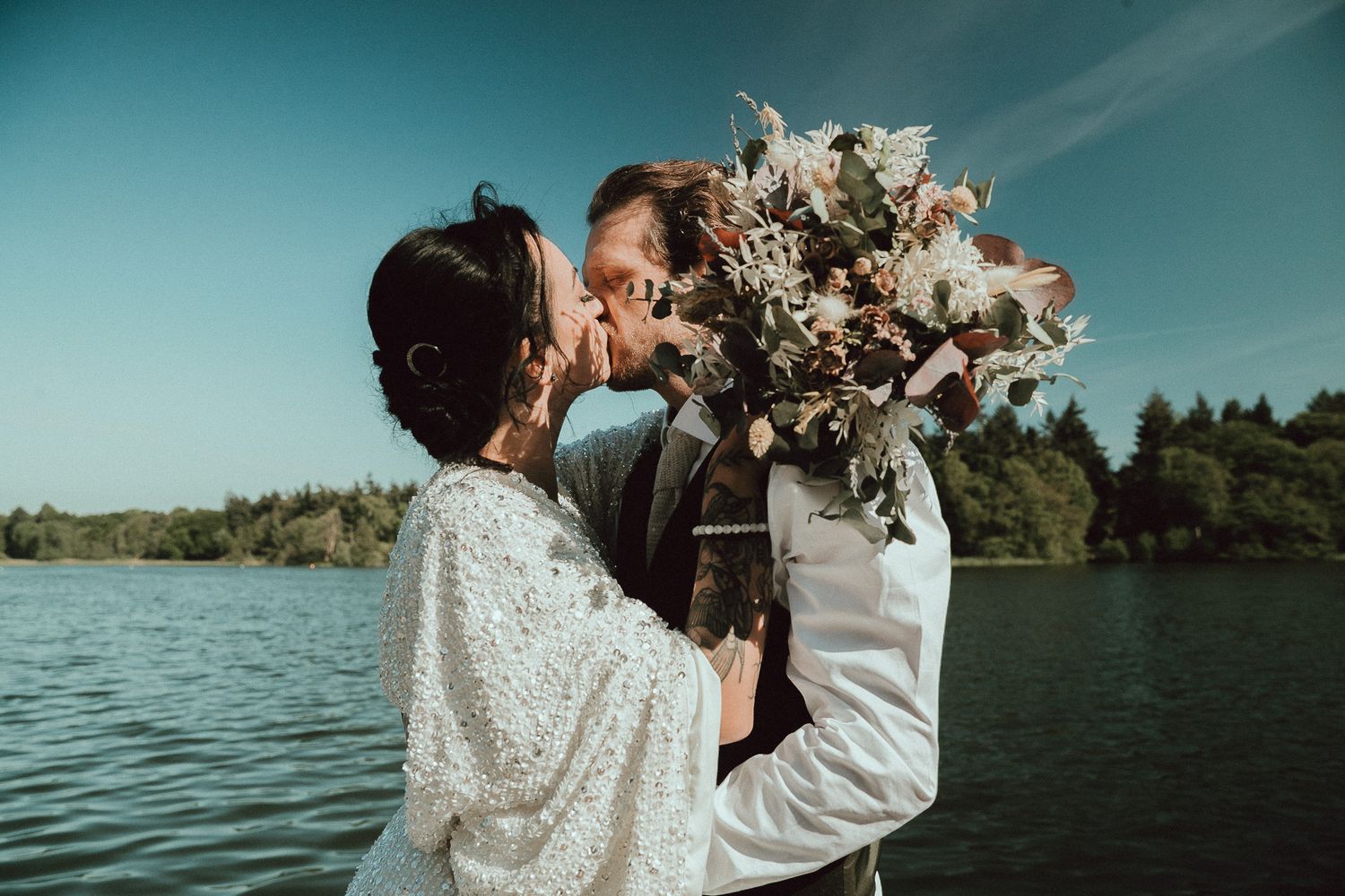 Couple embraces by a serene lake with mountains and trees in the background during a romantic outdoor moment.