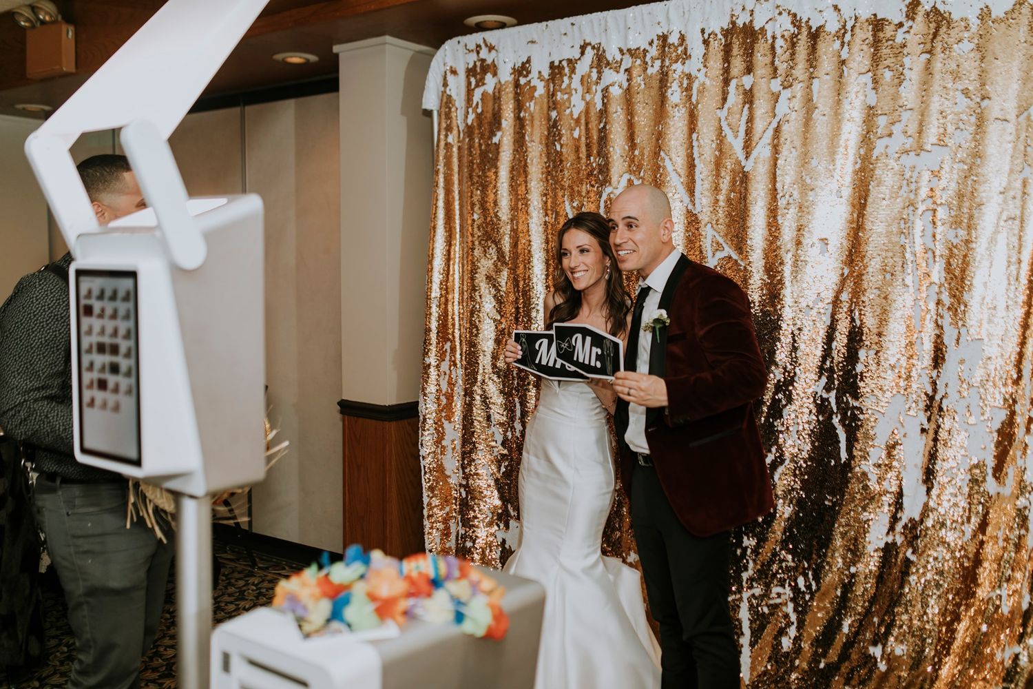 A couple stands in front of a sparkly gold sequin backdrop during an indoor ceremony with a white photo booth nearby.