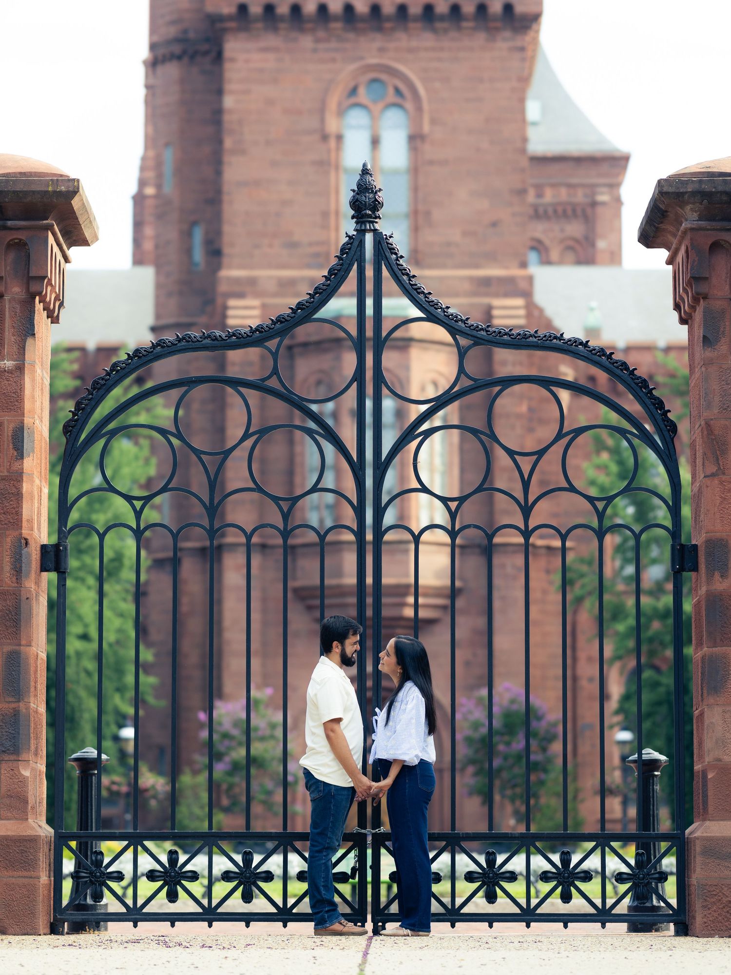 A couple stands together near ornate black iron gates at a historic red brick building with gothic architecture.