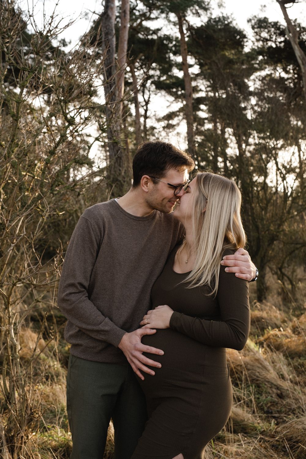 A couple in dark clothing shares an intimate moment outdoors among trees at sunset.