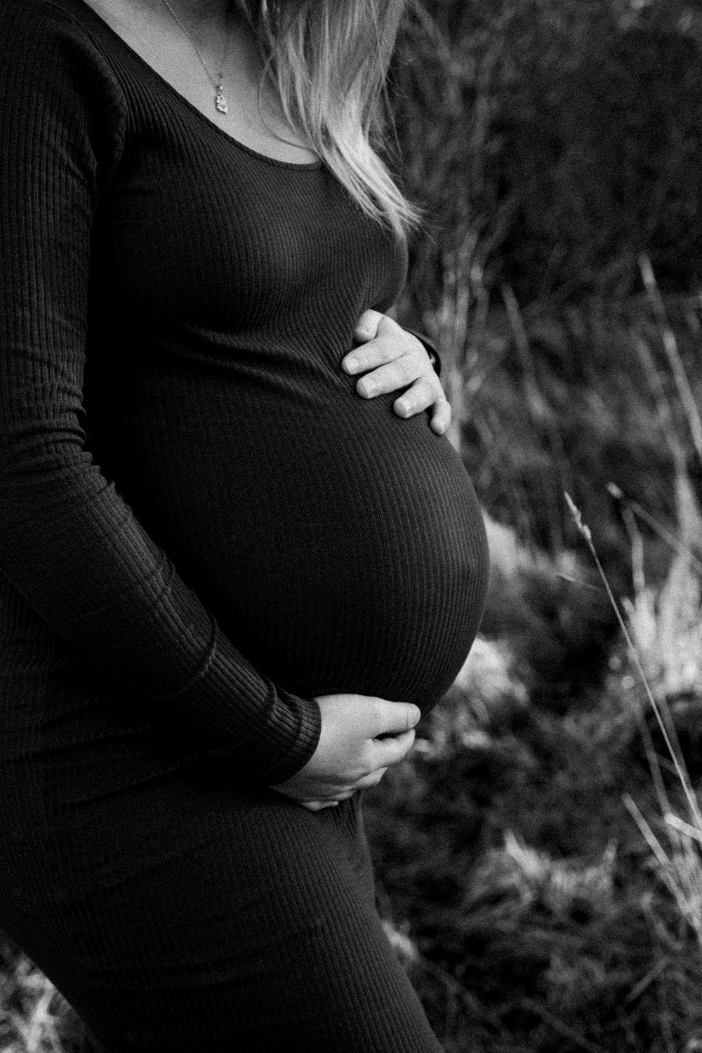 A black and white maternity photo showing a pregnant belly in a fitted black sweater against a natural outdoor background.