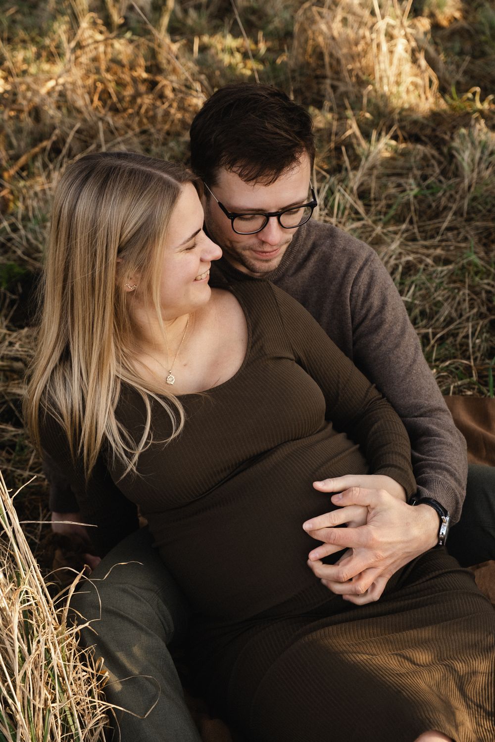 Expecting couple sits together in tall grass during golden hour sunset for maternity photo session.