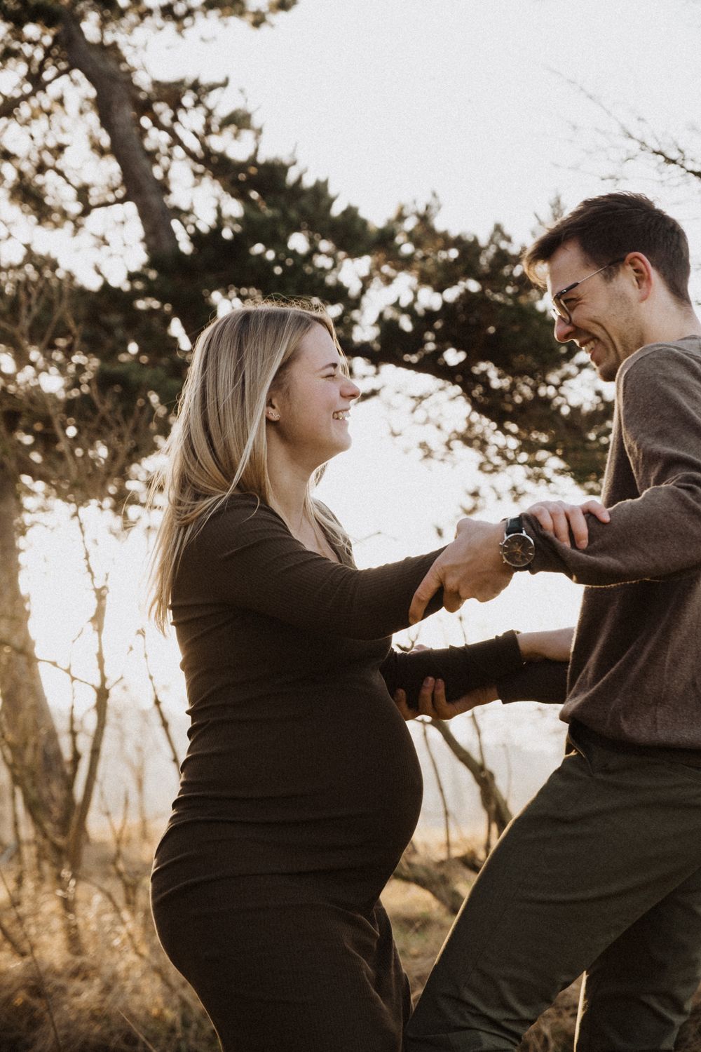 A couple shares a joyful moment dancing together outdoors at sunset, with the woman wearing a black maternity dress.