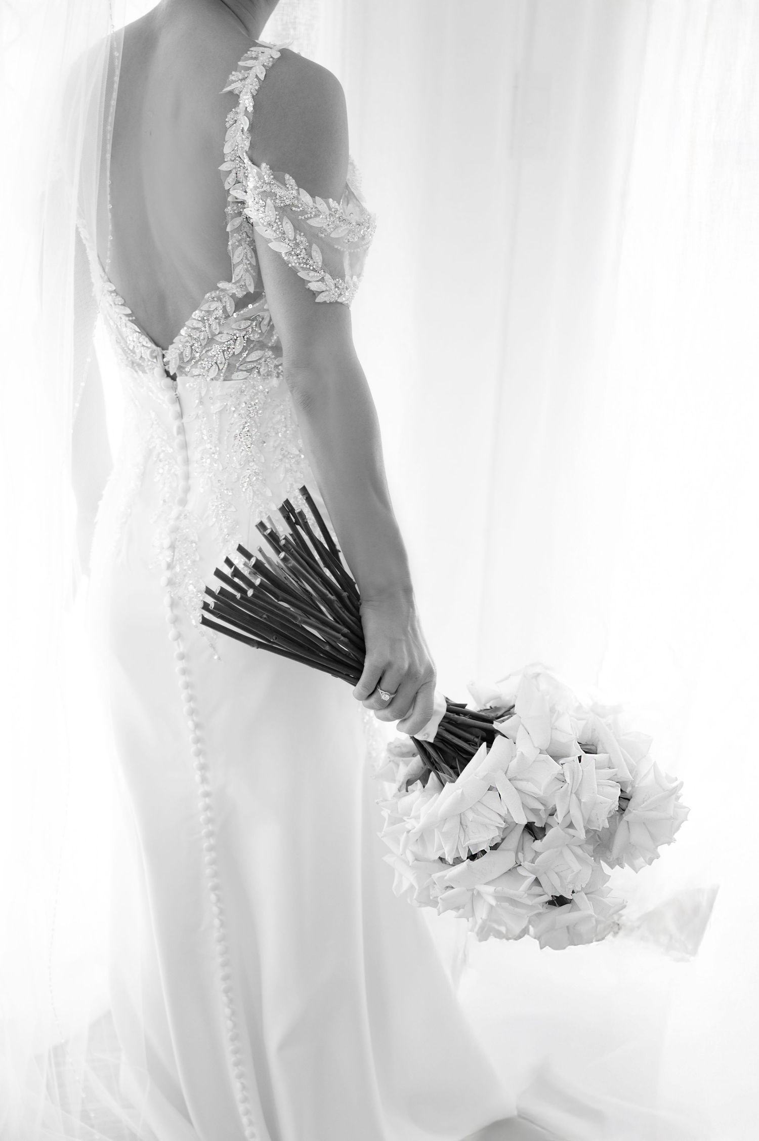 Elegant bride in backless lace wedding gown holding white rose bouquet by sheer curtains.