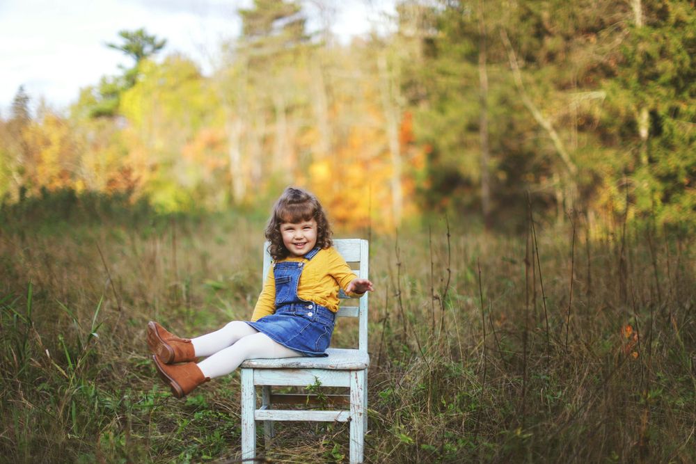 A person lounges on a white wooden chair reading a book in a field of tall grass during autumn.