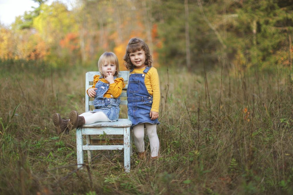 Two children in matching denim outfits sit together on a white wooden bench in an autumn field setting.