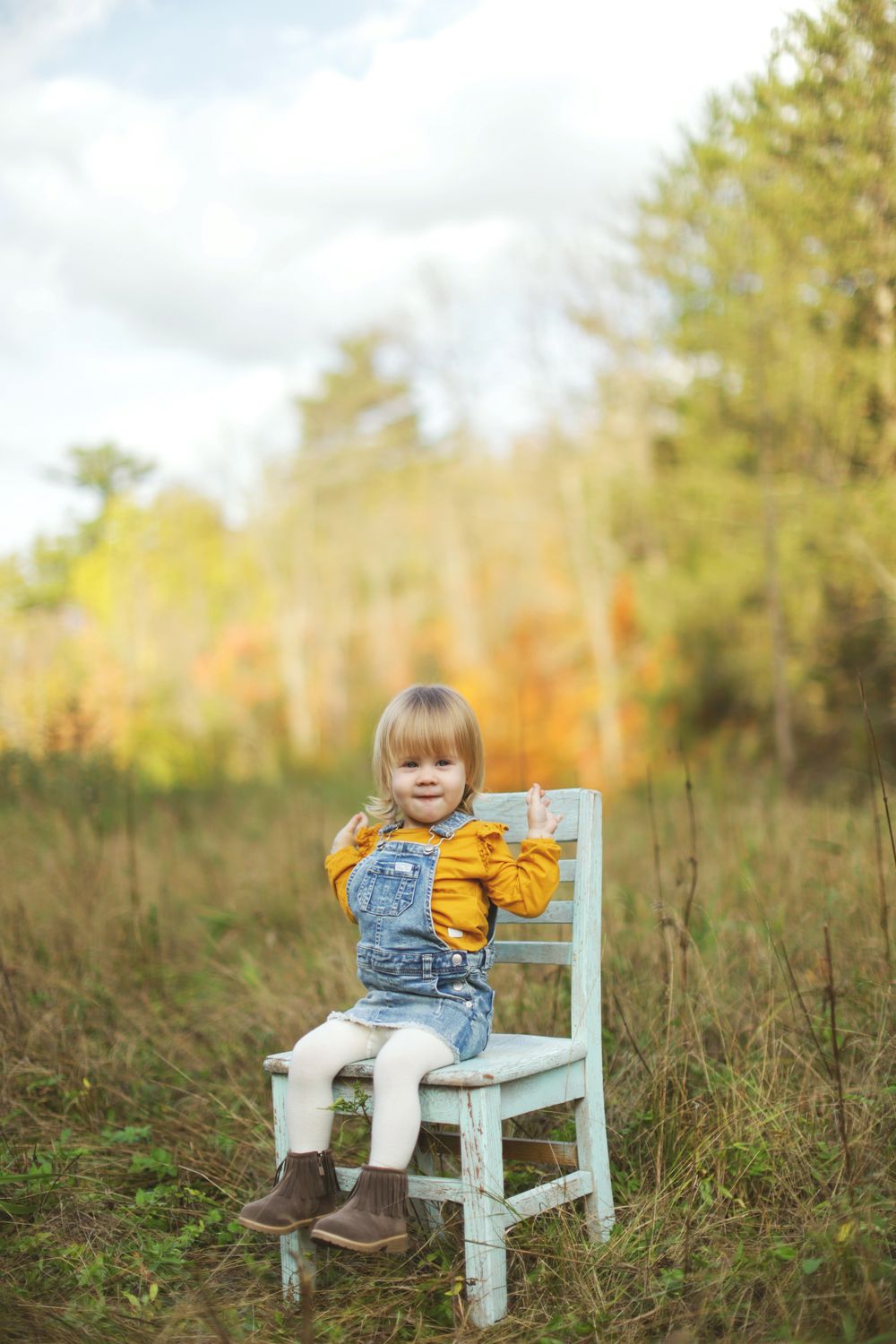 A small figure sits on a light blue wooden chair in a grassy field with autumn trees in the background.