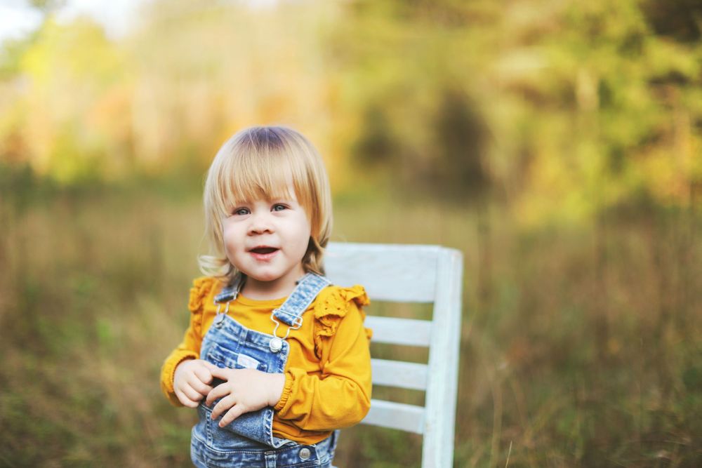 Child in yellow sweater sits on white chair surrounded by autumn foliage in outdoor photo session.
