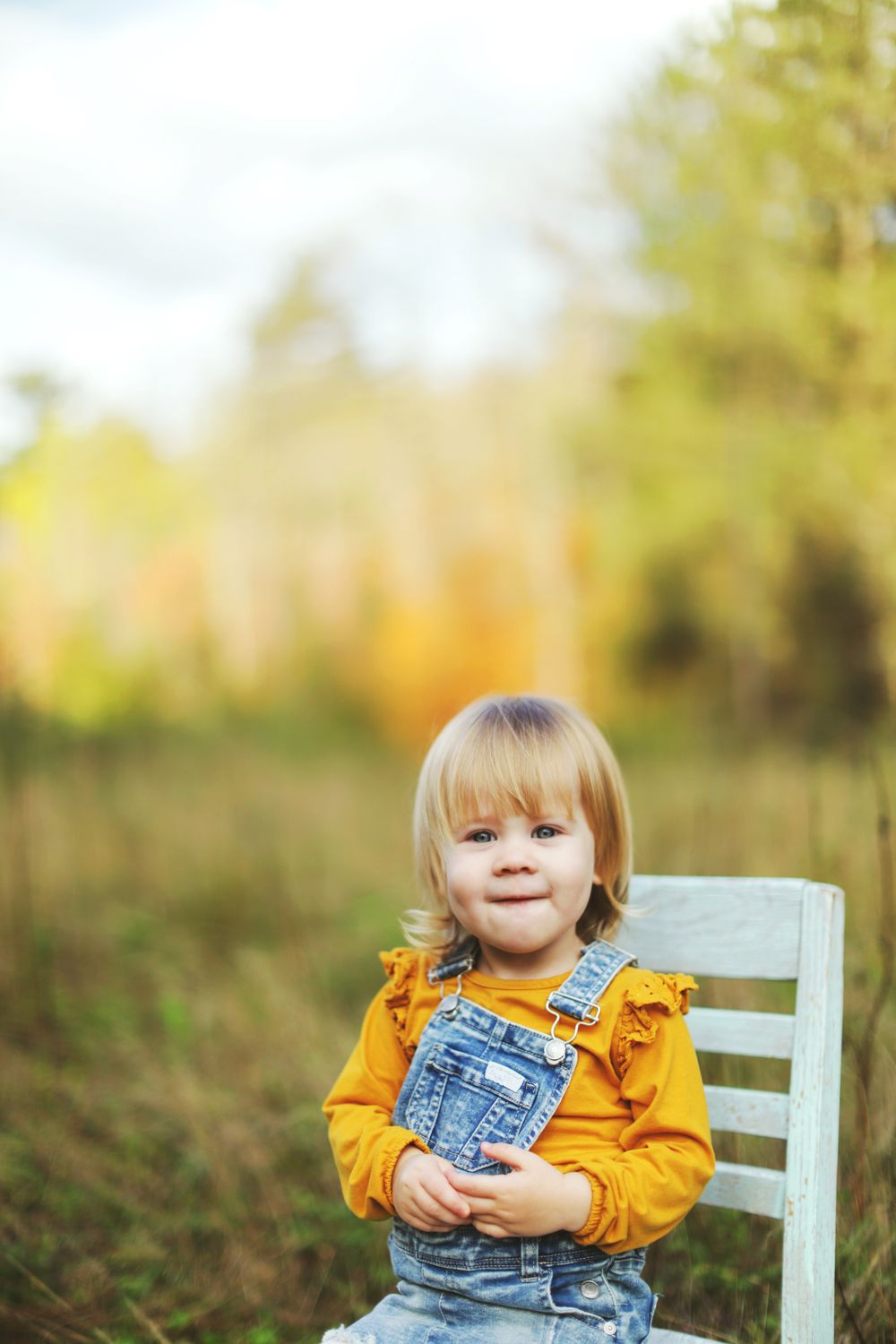 Child wearing yellow top and denim overalls sitting on a light blue chair outdoors.