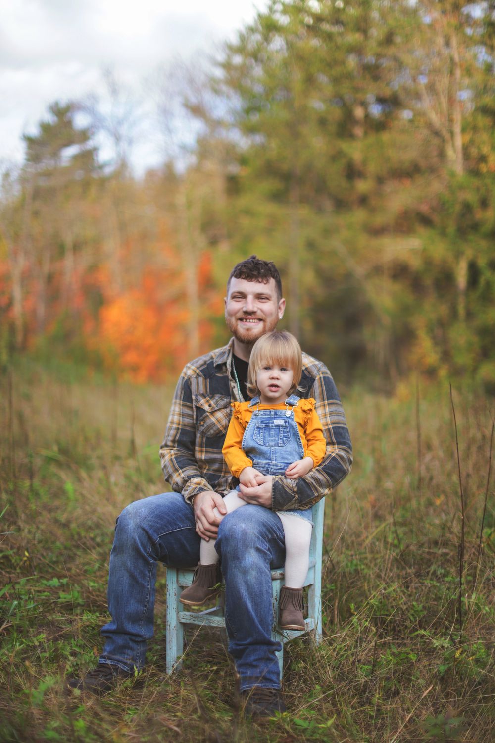 Person sitting in a blue vintage chair outdoors surrounded by tall grass and autumn trees.