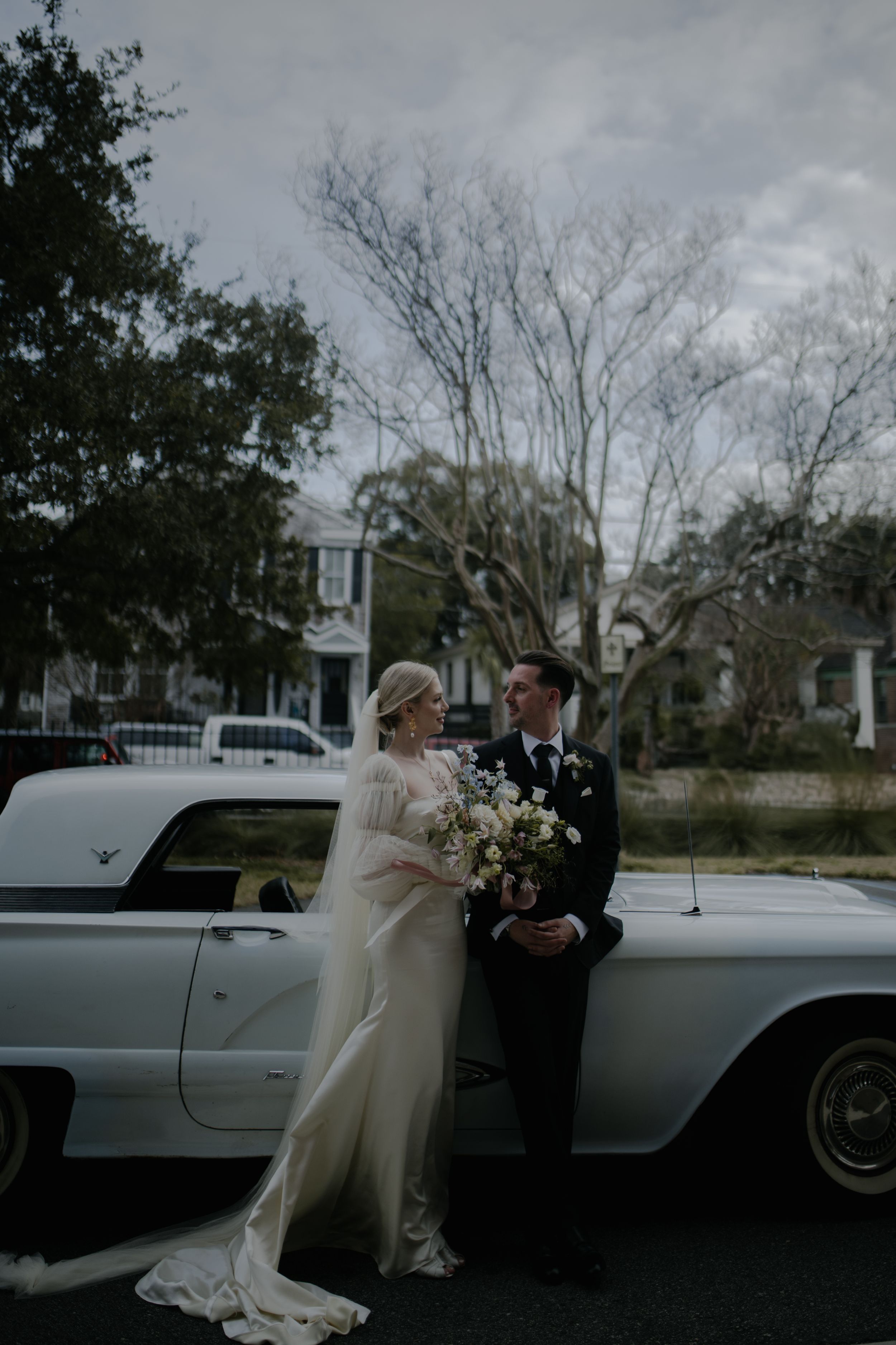 A romantic moment unfolds in front of a vintage car on a cloudy day with bare trees in the background.