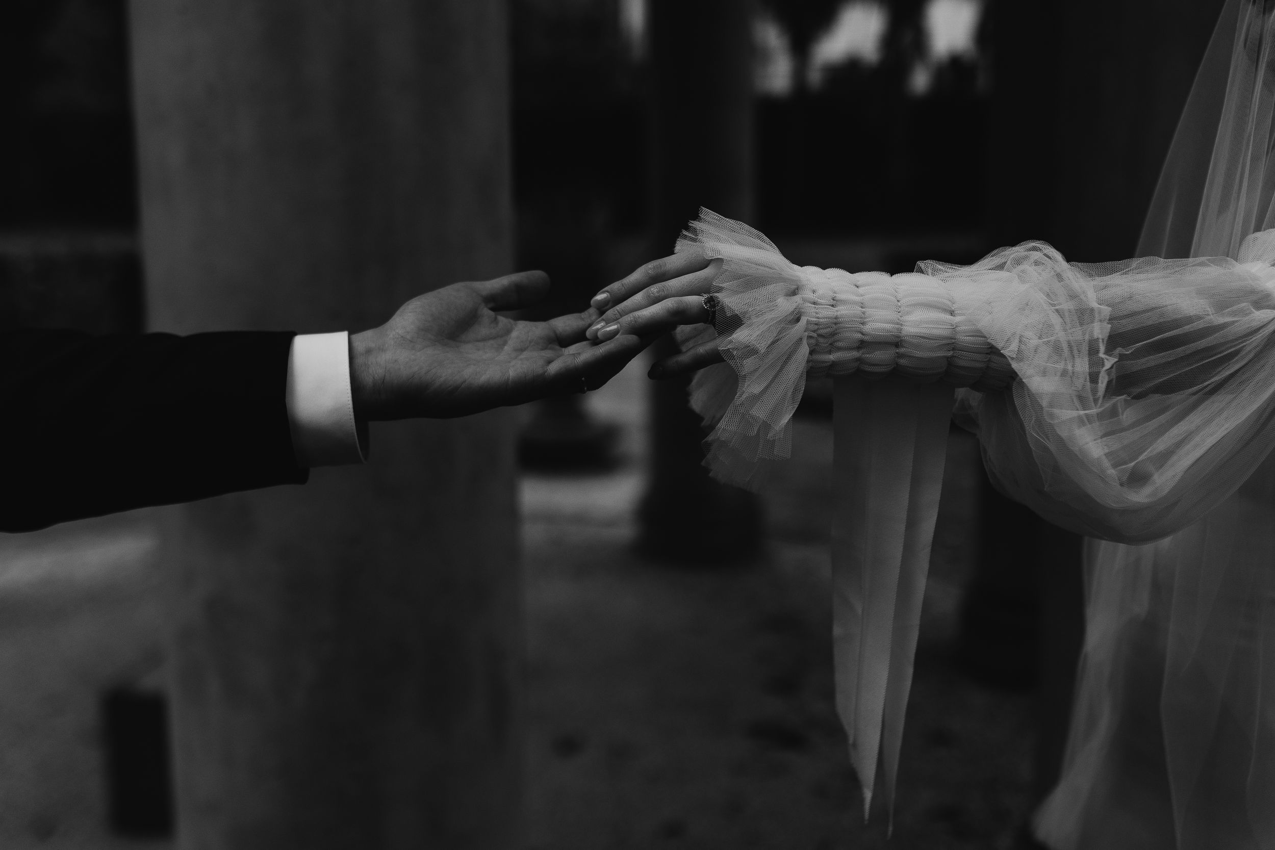 A black and white close-up photo of hands reaching for each other through delicate wedding veil fabric.