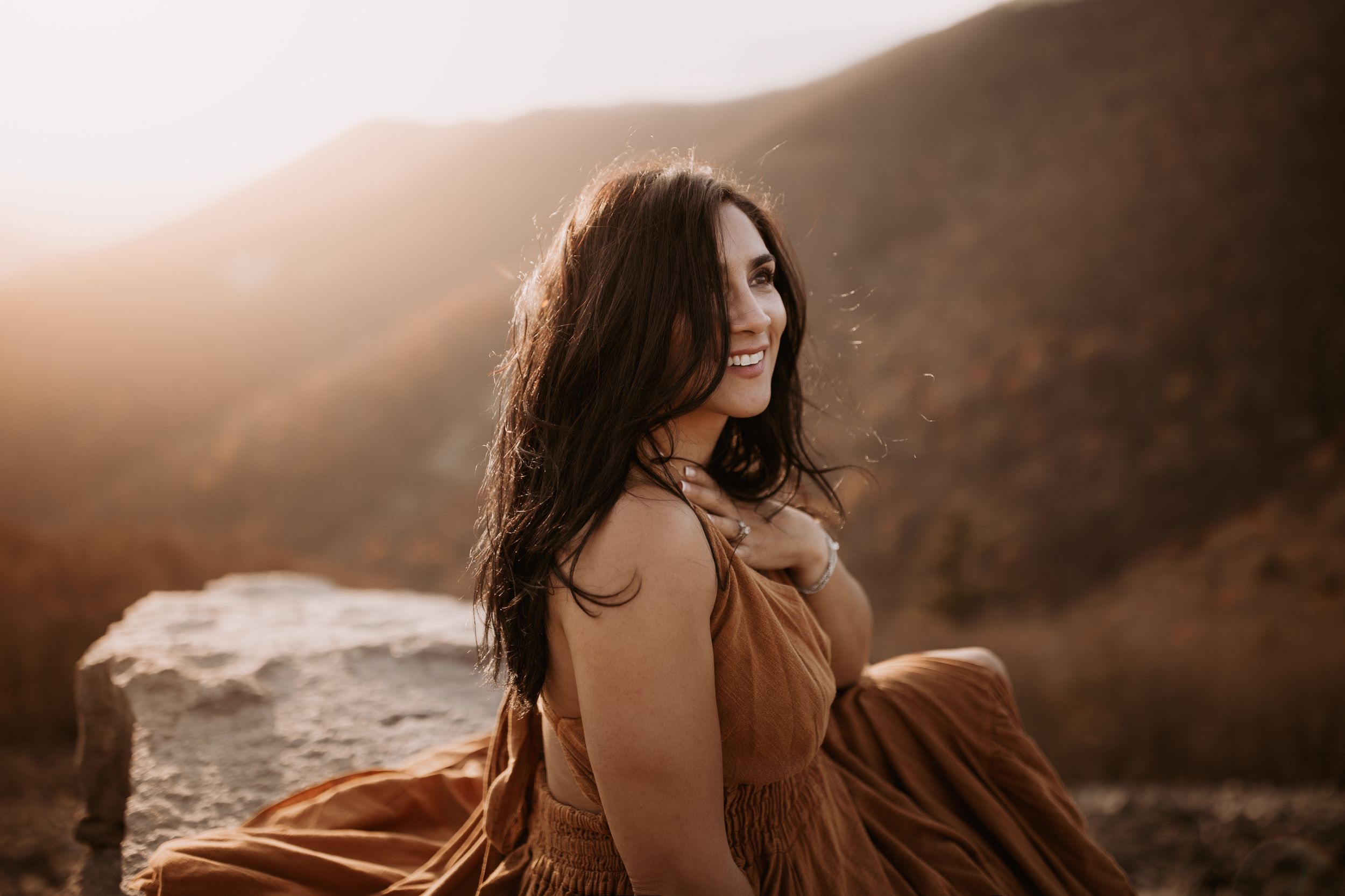 A woman in a flowing dress poses against a dramatic mountainous landscape at sunset.