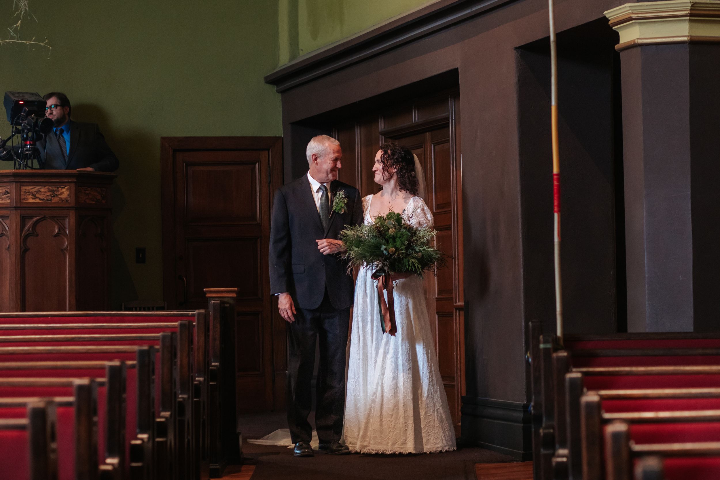 A couple stands in a dimly lit church aisle with red carpeting and ornate wooden pews.