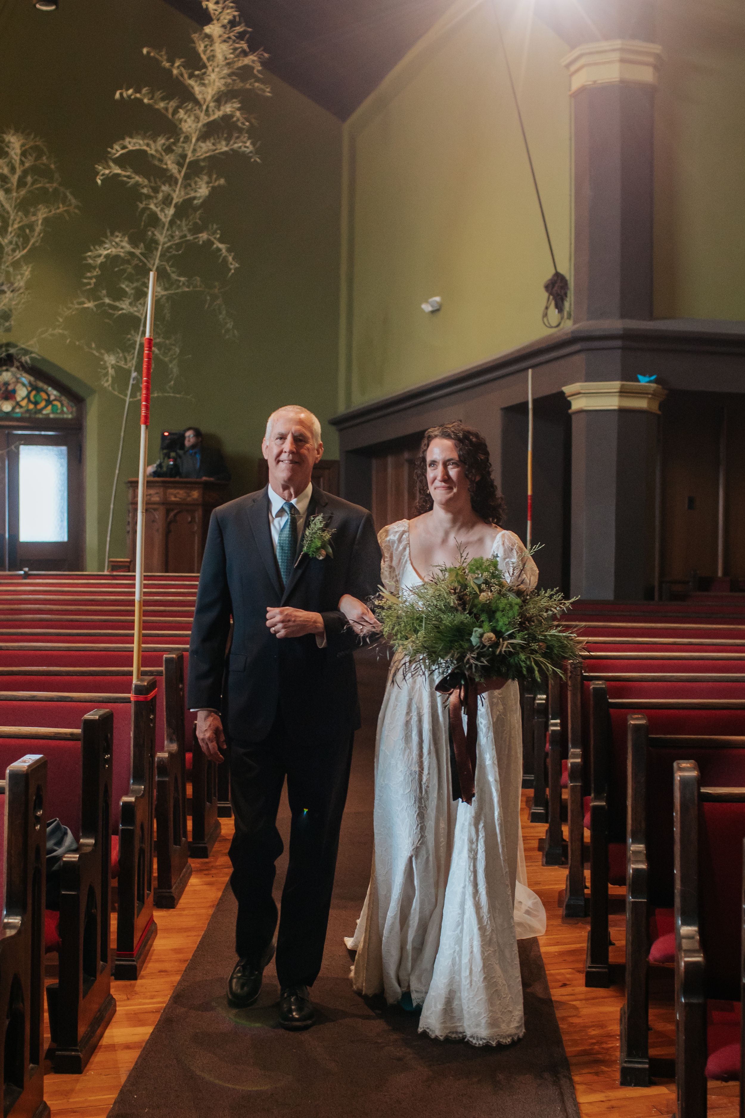A couple walks down the aisle of a traditional church with wooden pews and sunlight streaming through the windows.