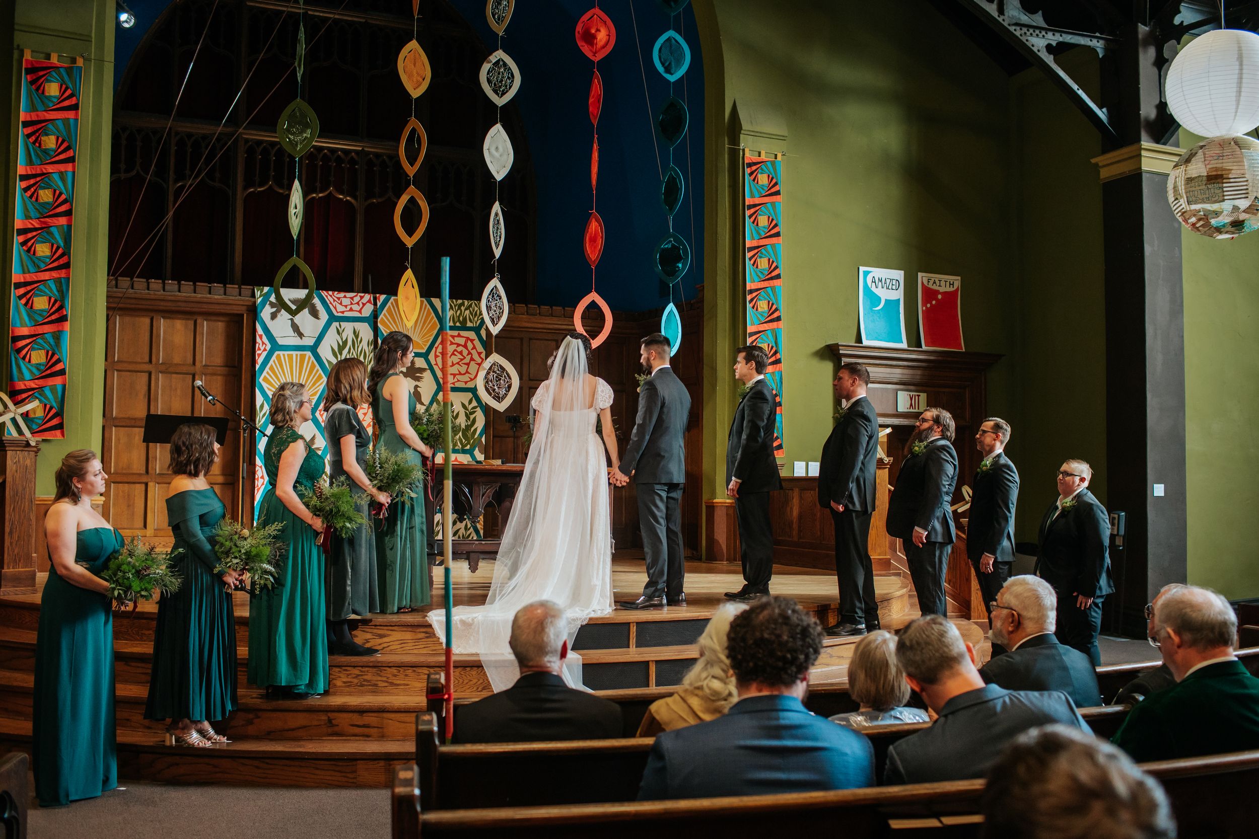 Colorful decorative hanging ornaments adorn a wedding ceremony venue with wooden walls and green accents.
