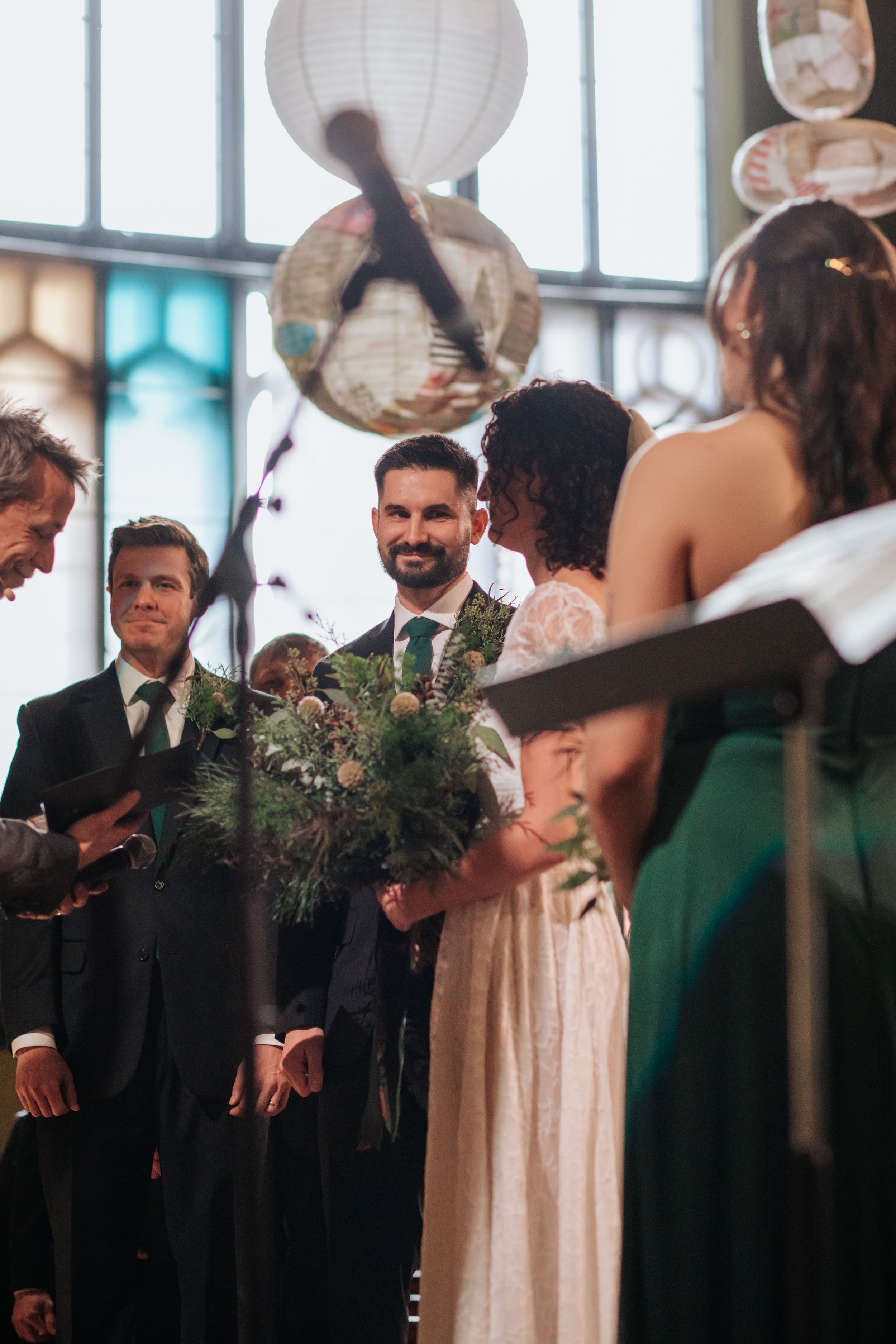 Wedding party members stand in a line during an indoor ceremony with natural lighting streaming through windows.