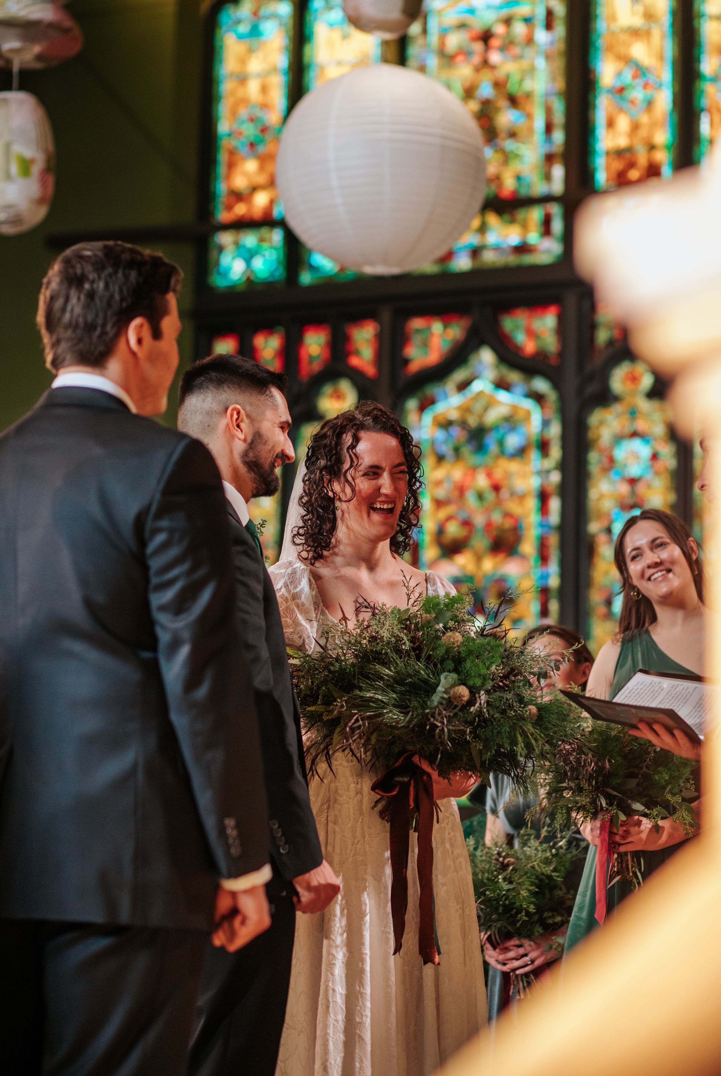 A wedding ceremony takes place in a church with stained glass windows and paper lanterns, featuring a lush evergreen bouquet.