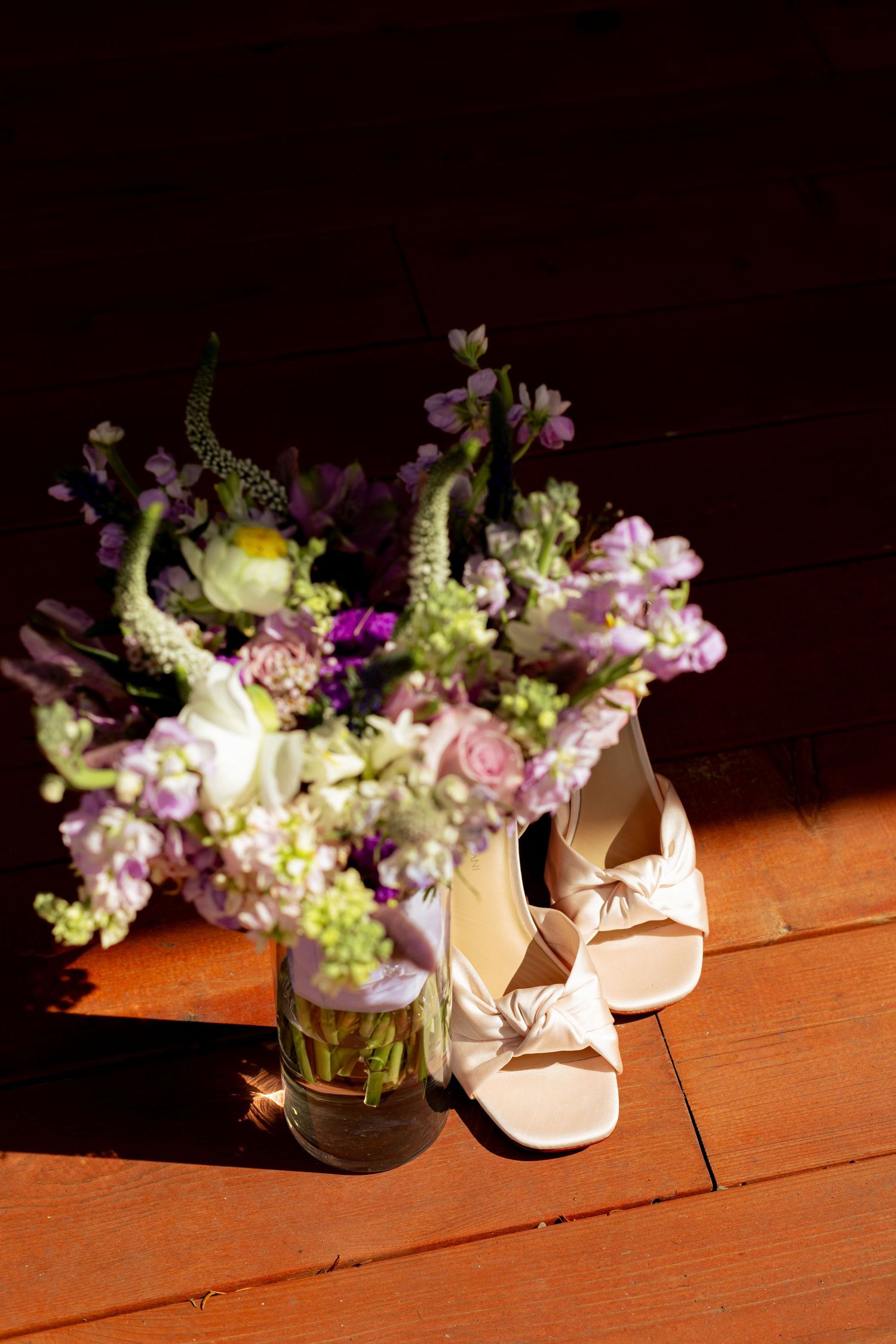 Pink and white flowers arranged in glass vases next to beige shoes on a wooden surface in dramatic lighting.