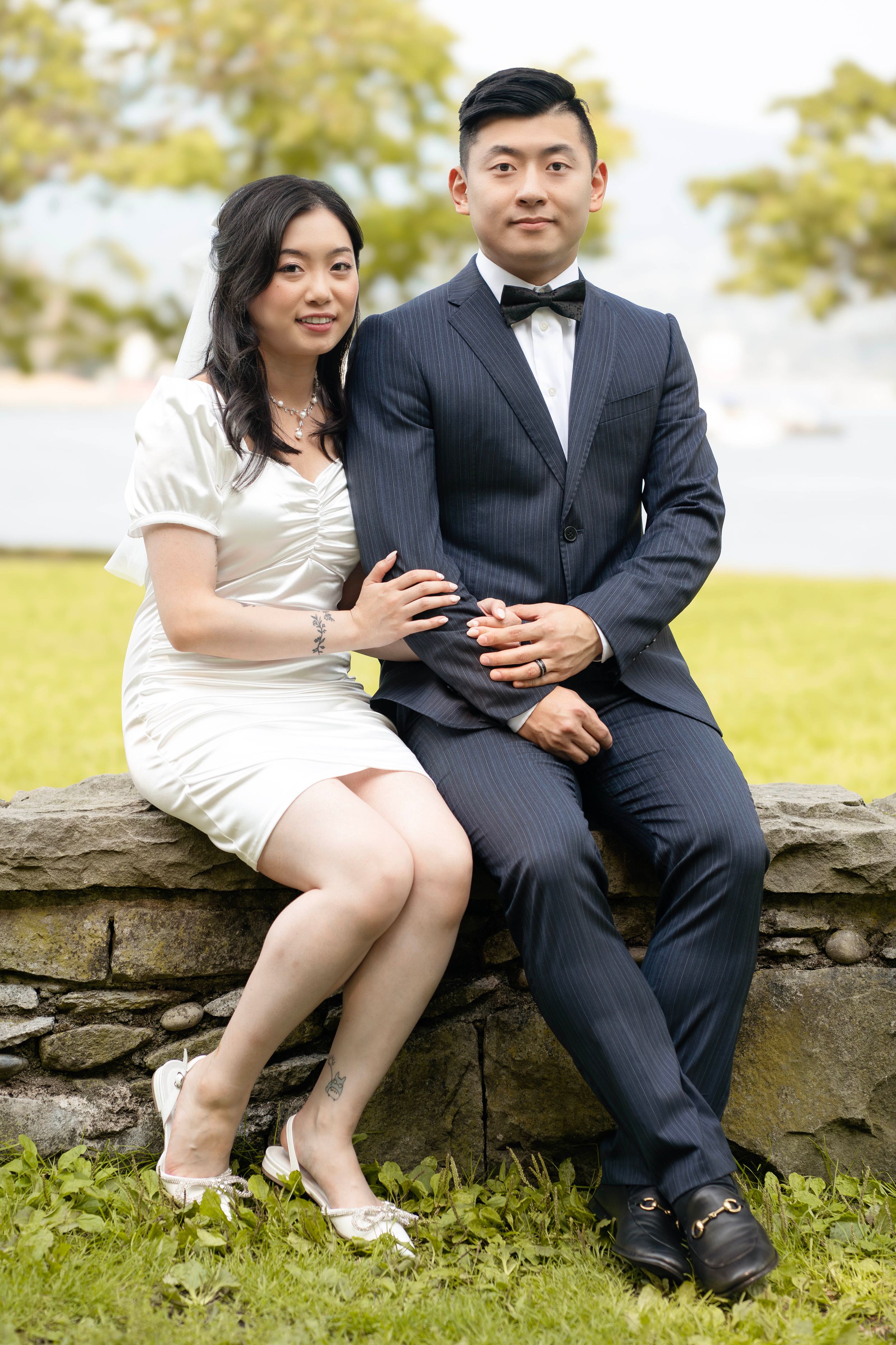 A couple sits together on a stone wall outdoors, dressed in formal attire with a white dress and dark suit.