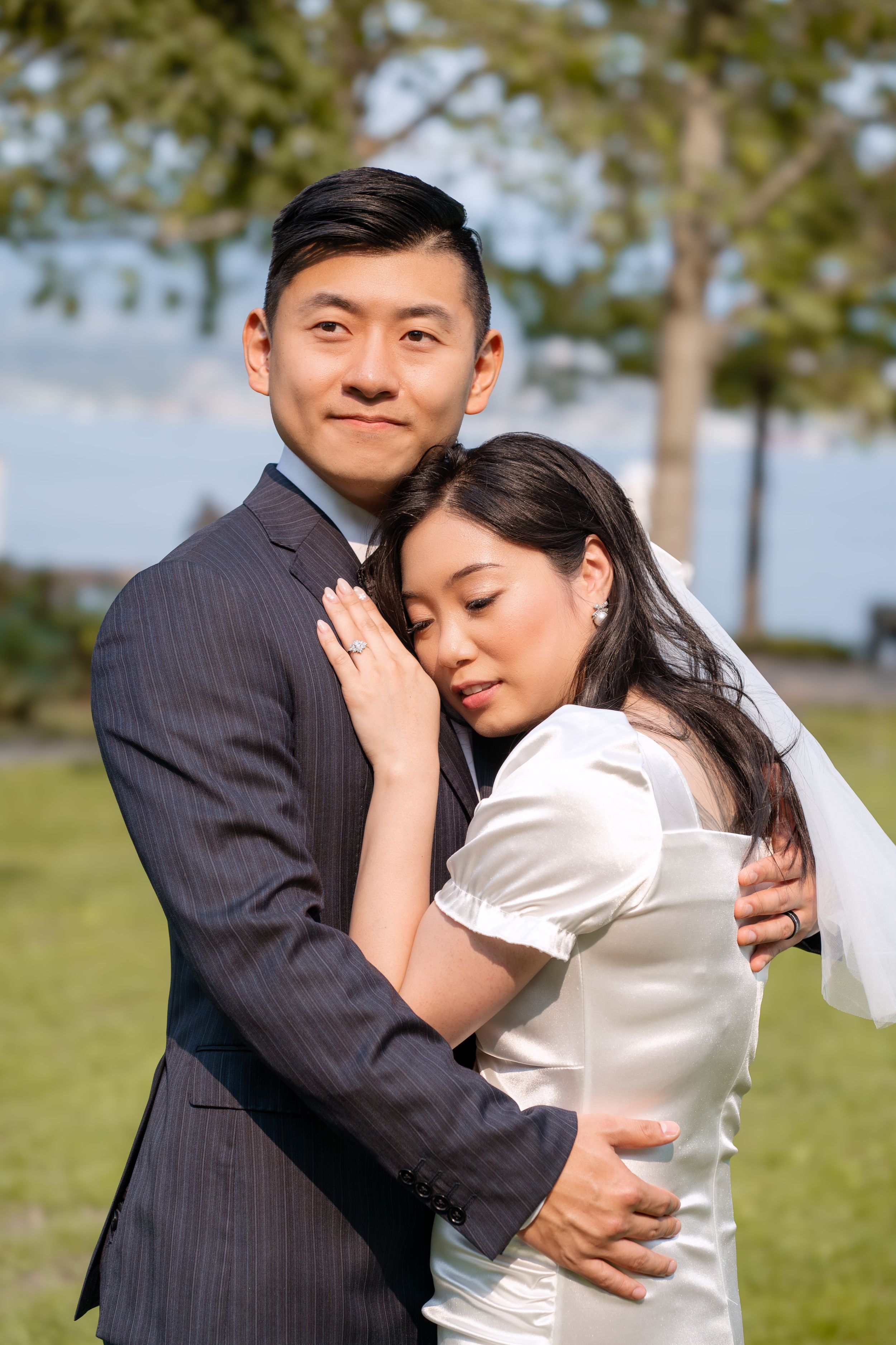 A couple embraces tenderly in a sunny outdoor setting with green grass and trees in the background.