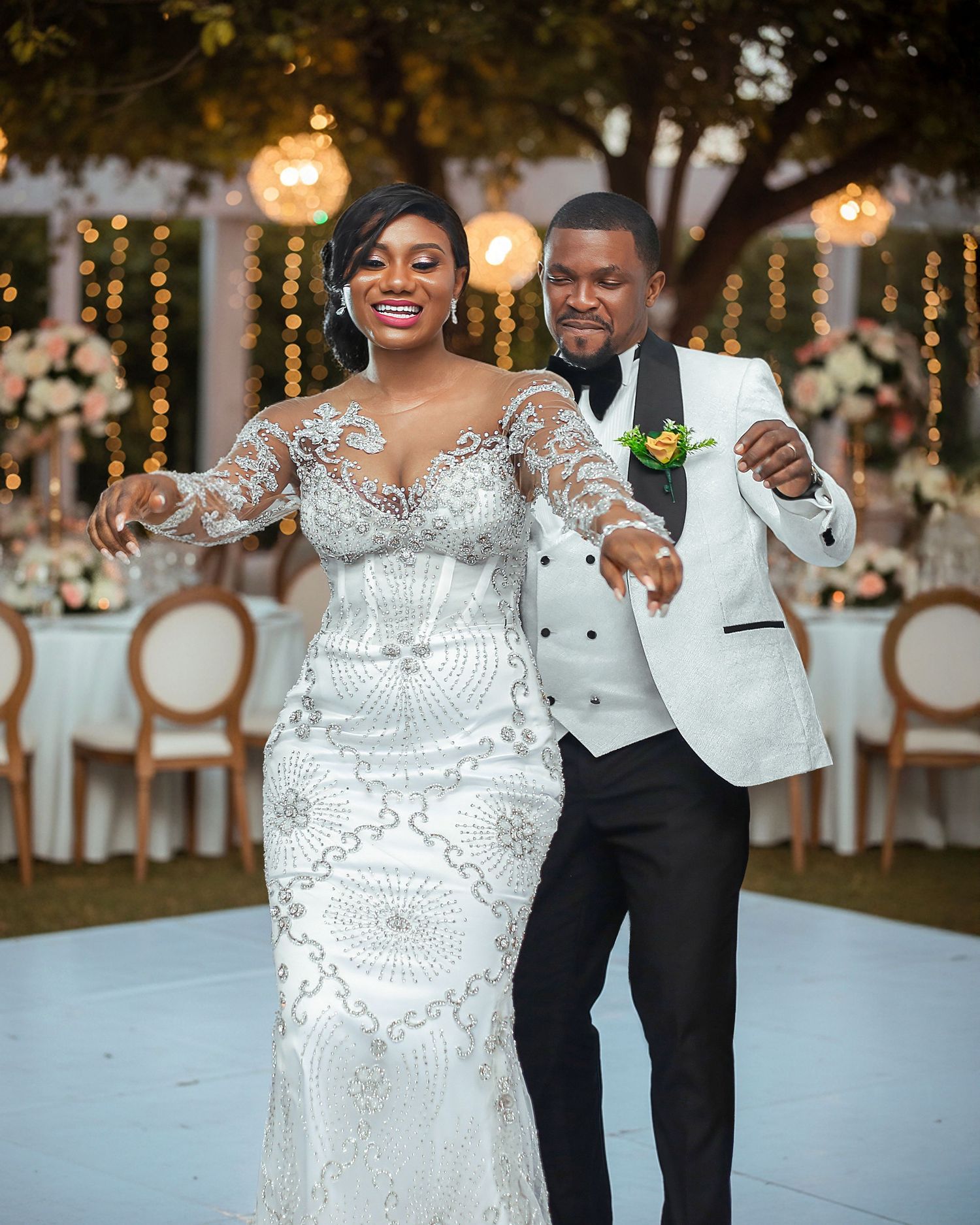 Bride and groom dancing joyfully at their wedding reception, bride in beaded gown, groom in white tuxedo jacket.