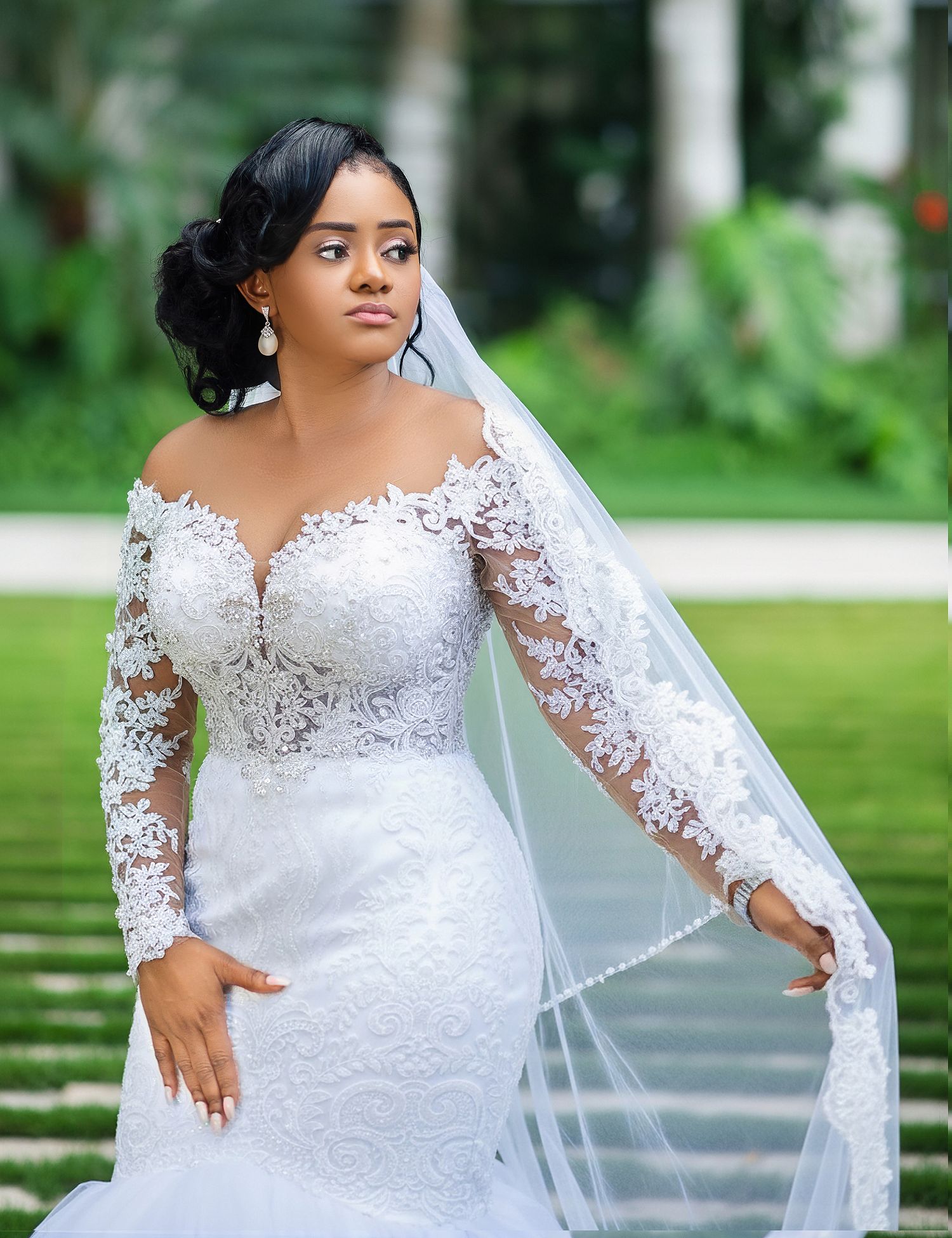 Bride in off-shoulder lace wedding gown with long sleeves and flowing veil poses outdoors amid lush greenery.
