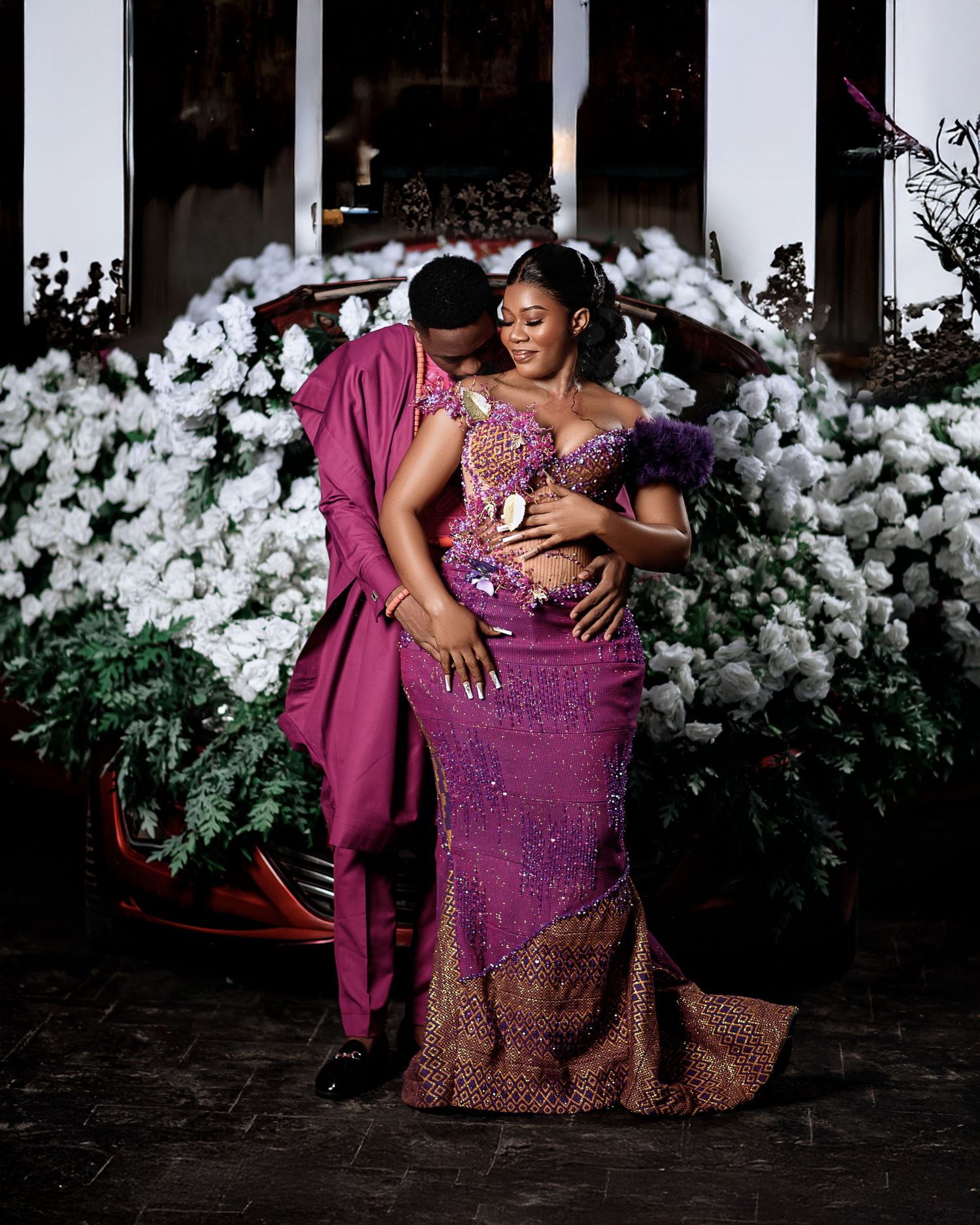 A couple in matching burgundy formal attire embrace in front of white floral decorations during a romantic photoshoot.