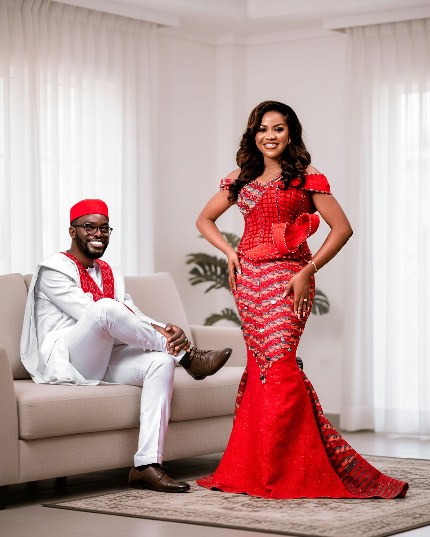 A couple in coordinated red and white traditional African attire pose elegantly in a bright modern living room setting.