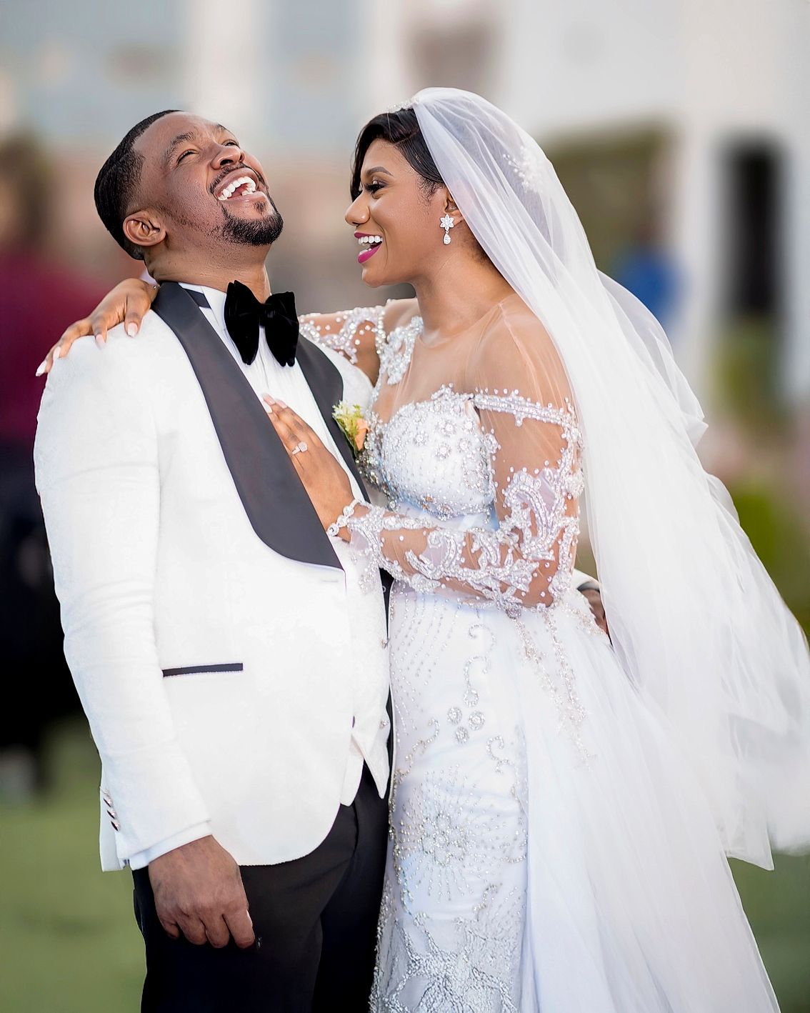 Joyful bride and groom in elegant white wedding attire laughing together outdoors on their wedding day.