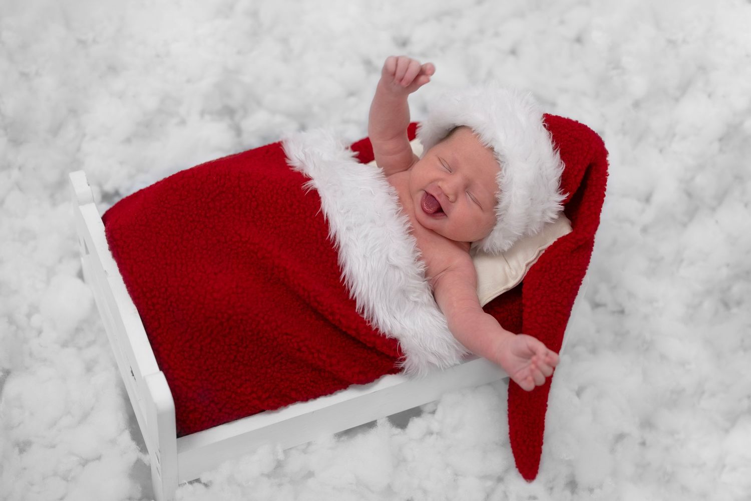 Newborn baby wearing Santa hat lying in small bed with tongue out during festive newborn photography session