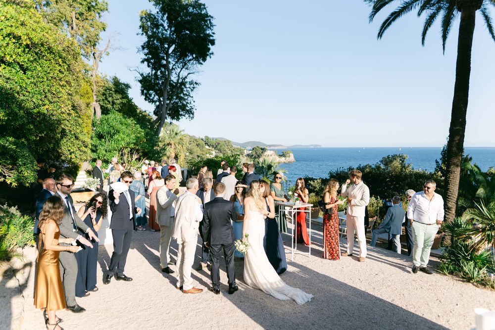 A group of guests gather at an elegant outdoor wedding ceremony with ocean views and palm trees in the background.