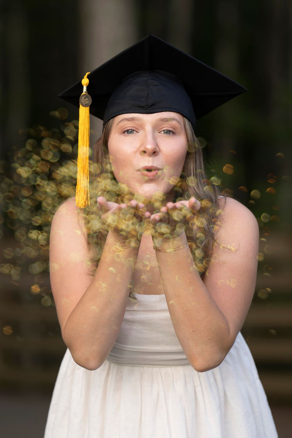 A graduate in a white dress and cap with gold tassel blows glitter from their hands in an outdoor celebration photo.