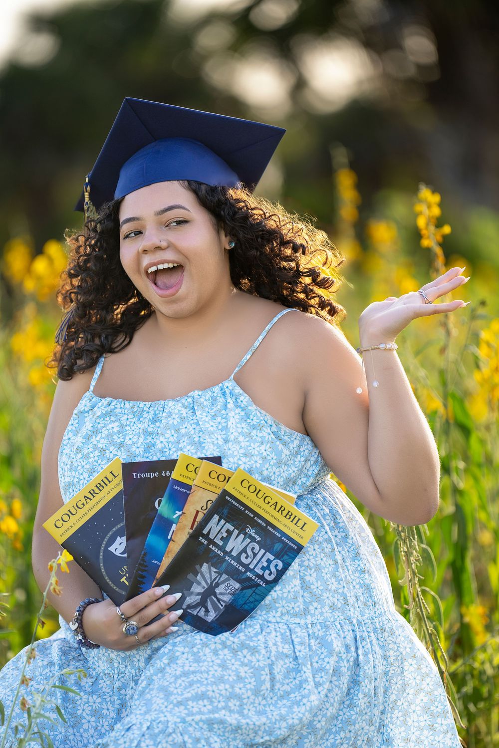 A person in a blue floral dress sits joyfully among yellow wildflowers while holding several books.