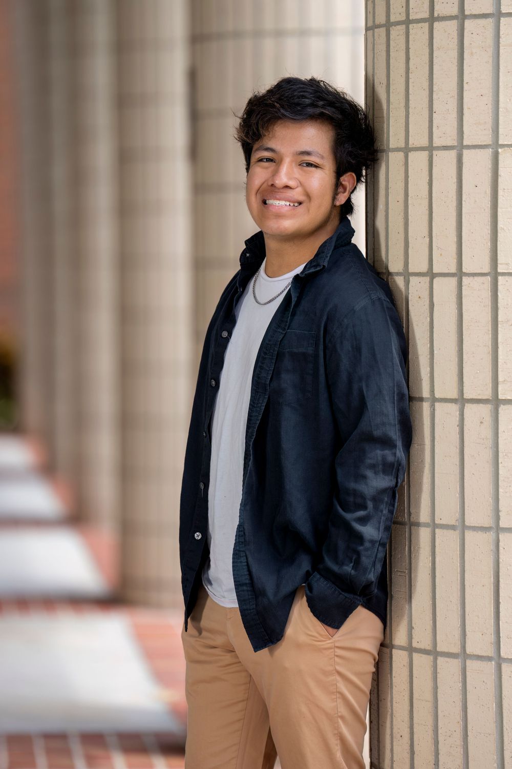 Young person in a navy jacket and khaki pants smiles while leaning against a beige wall.