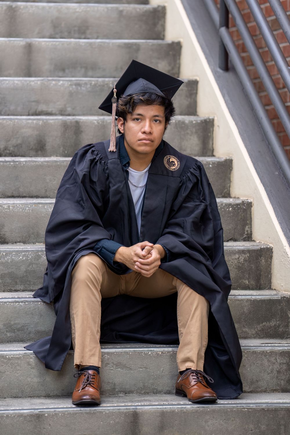Graduate in black cap and gown sits thoughtfully on concrete steps wearing khaki pants and brown dress shoes.