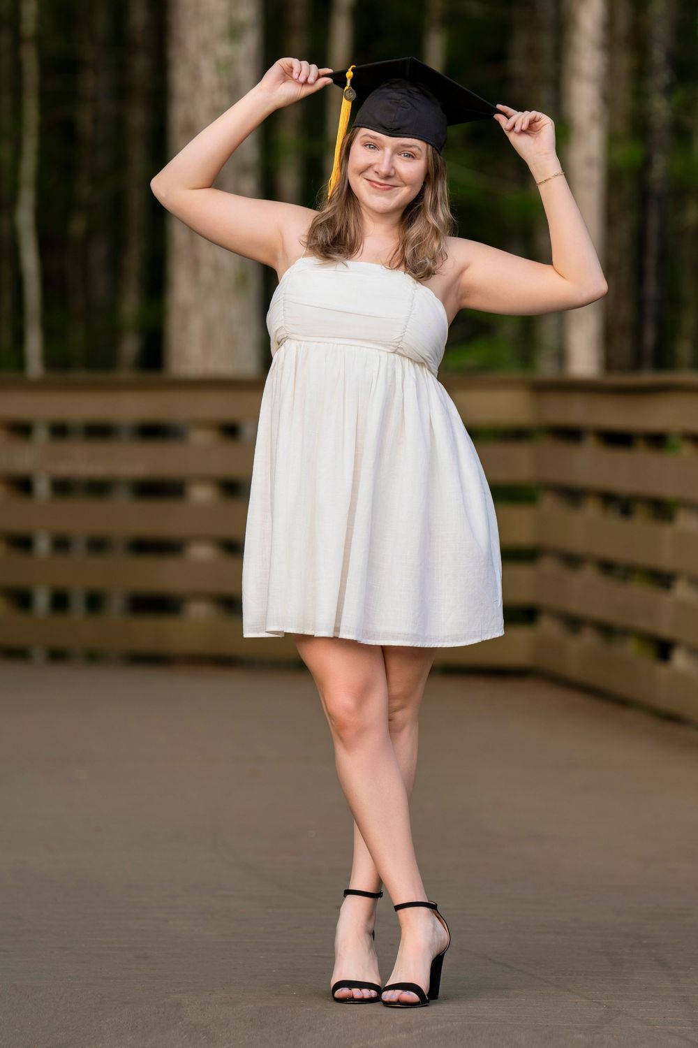 A graduate in a white dress and mortarboard cap poses playfully on a wooden deck surrounded by trees.
