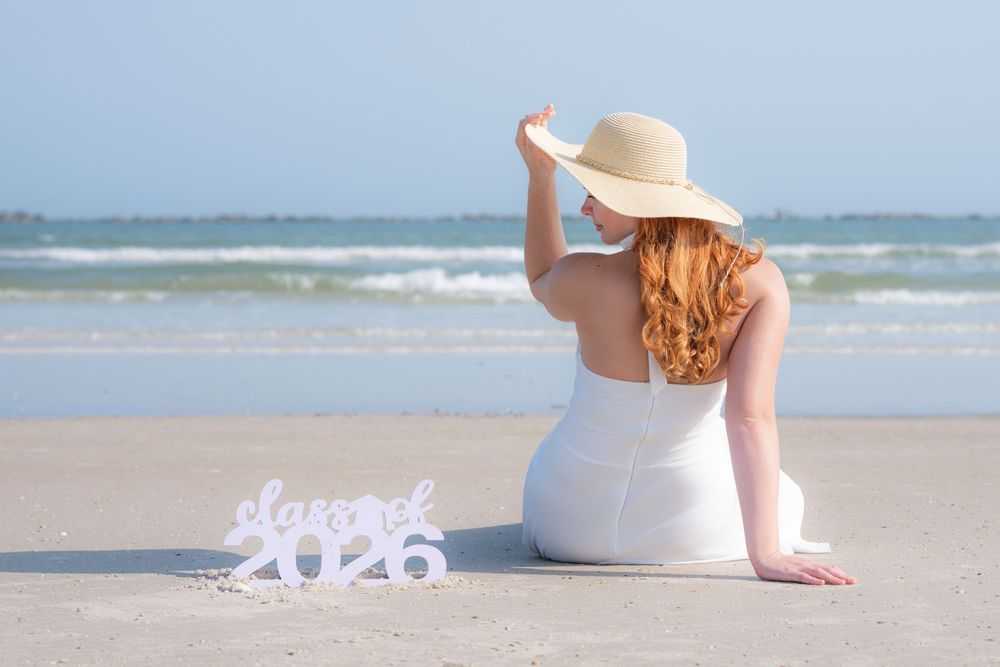 Someone in a white dress and sun hat sits on a sandy beach with ocean waves in the background.