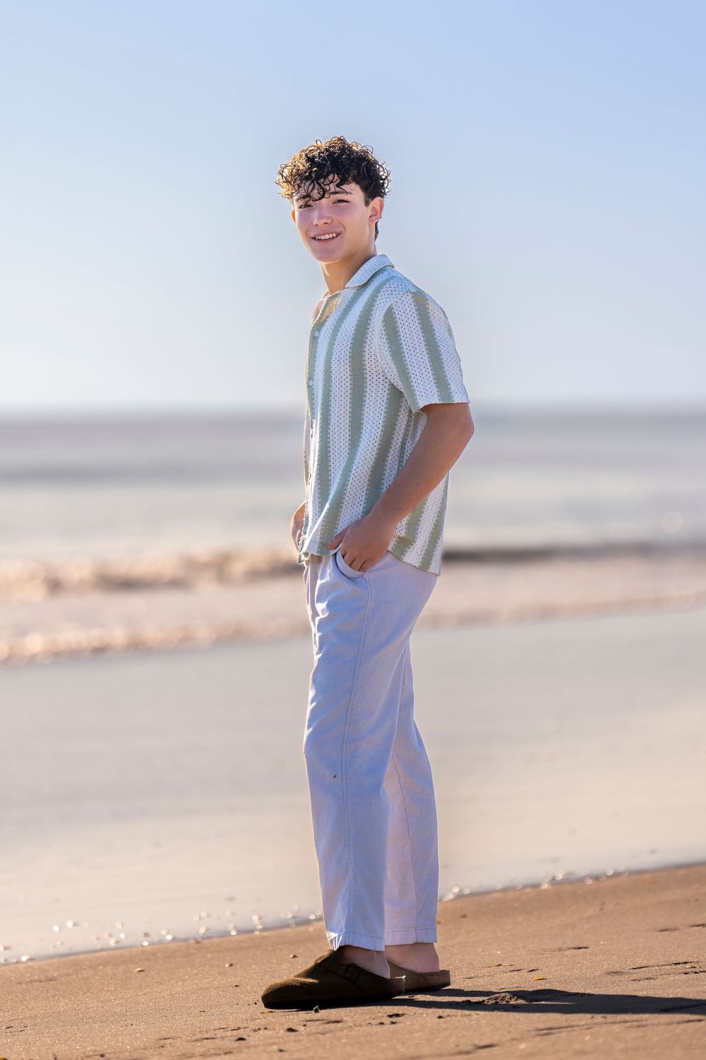 Someone in a striped shirt and light pants poses casually on a sandy beach during golden hour.