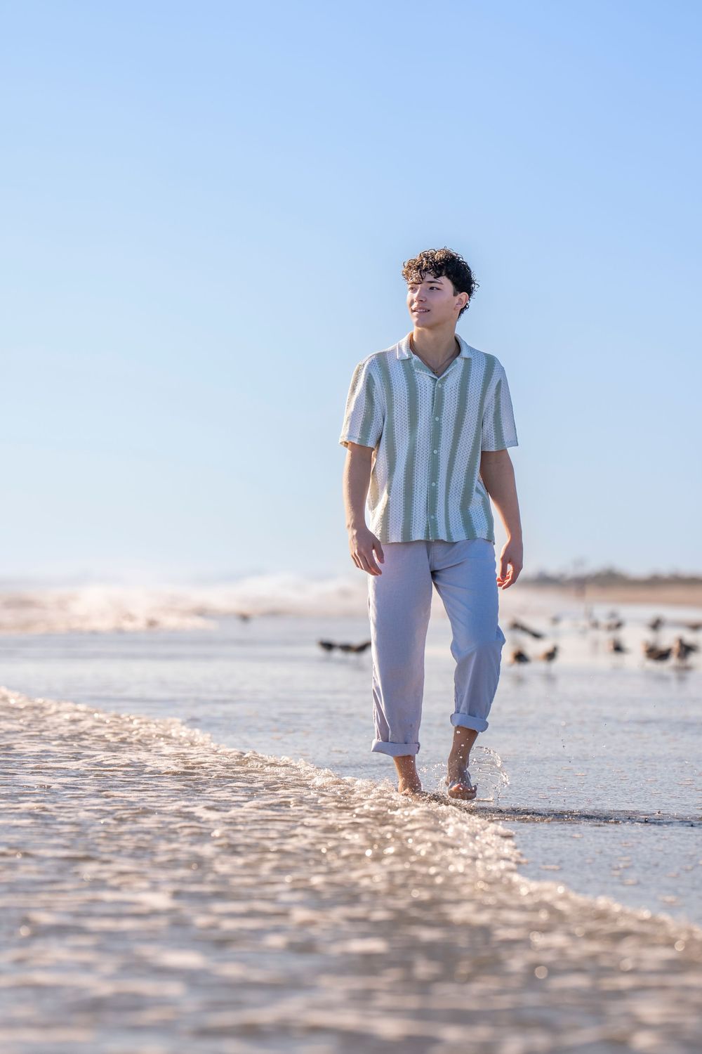 Someone wearing a striped shirt and rolled up pants walks barefoot along a sunny beach shoreline.