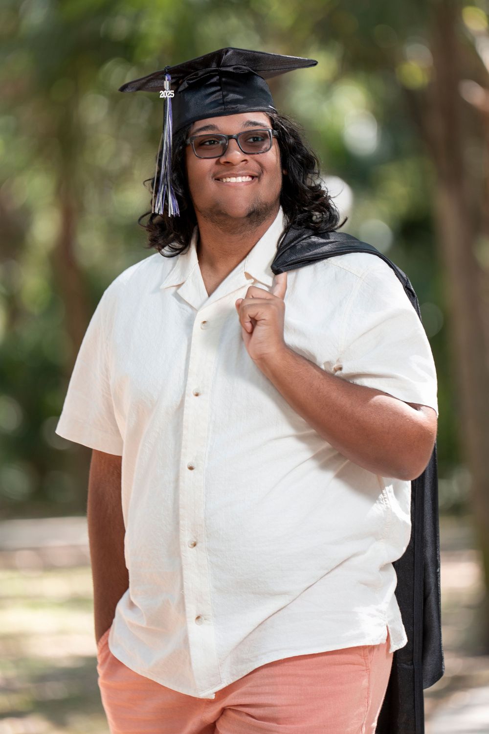 Person in a white button-down shirt and peach-colored pants poses outdoors in a natural setting.