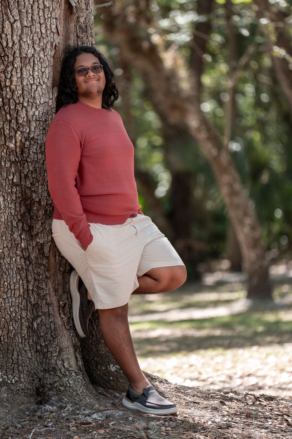 Person in coral sweater and white shorts leans casually against large tree trunk in outdoor setting.