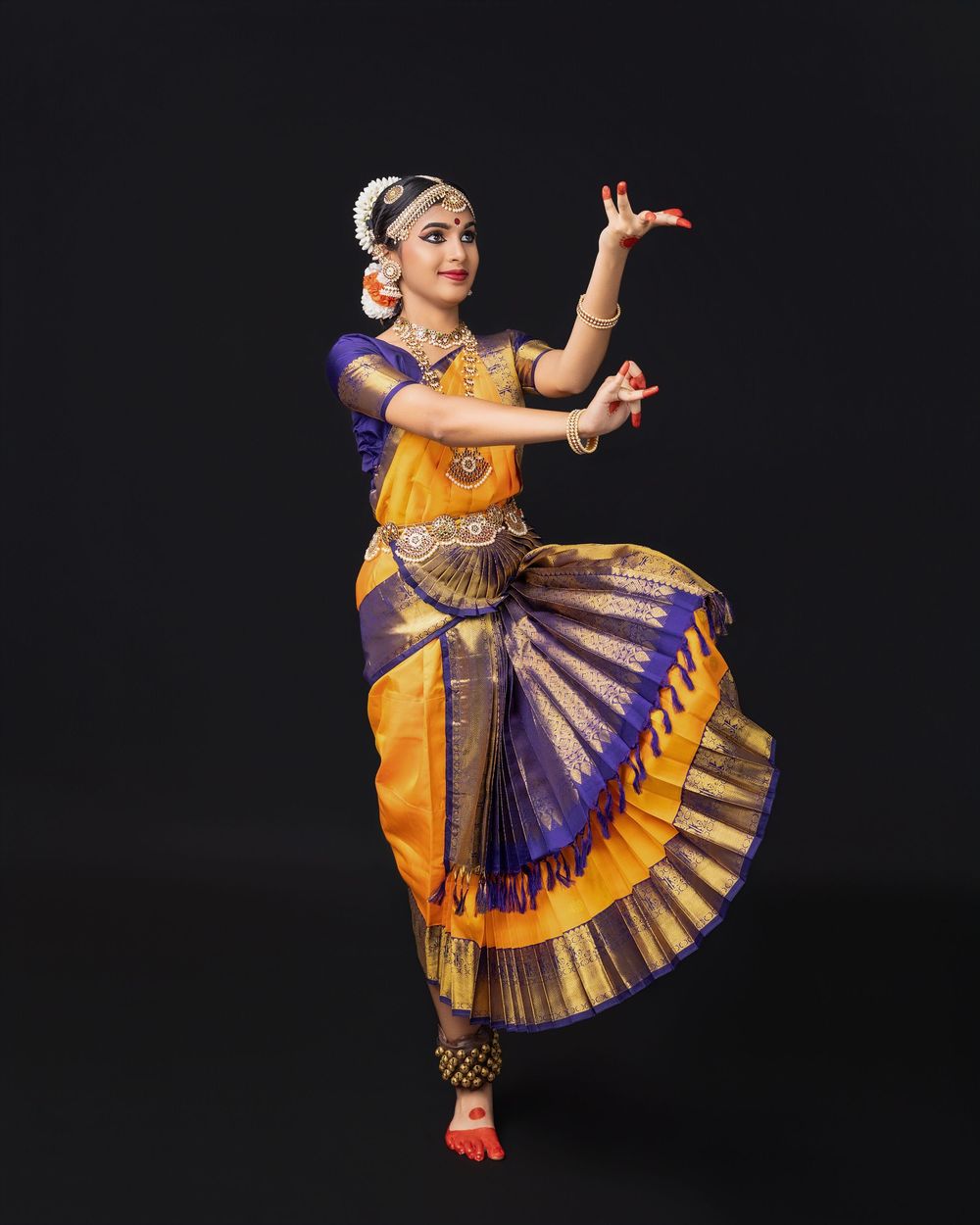 Classical Indian dancer in traditional yellow and purple attire performs expressive Bharatanatyam dance poses against black backdrop.