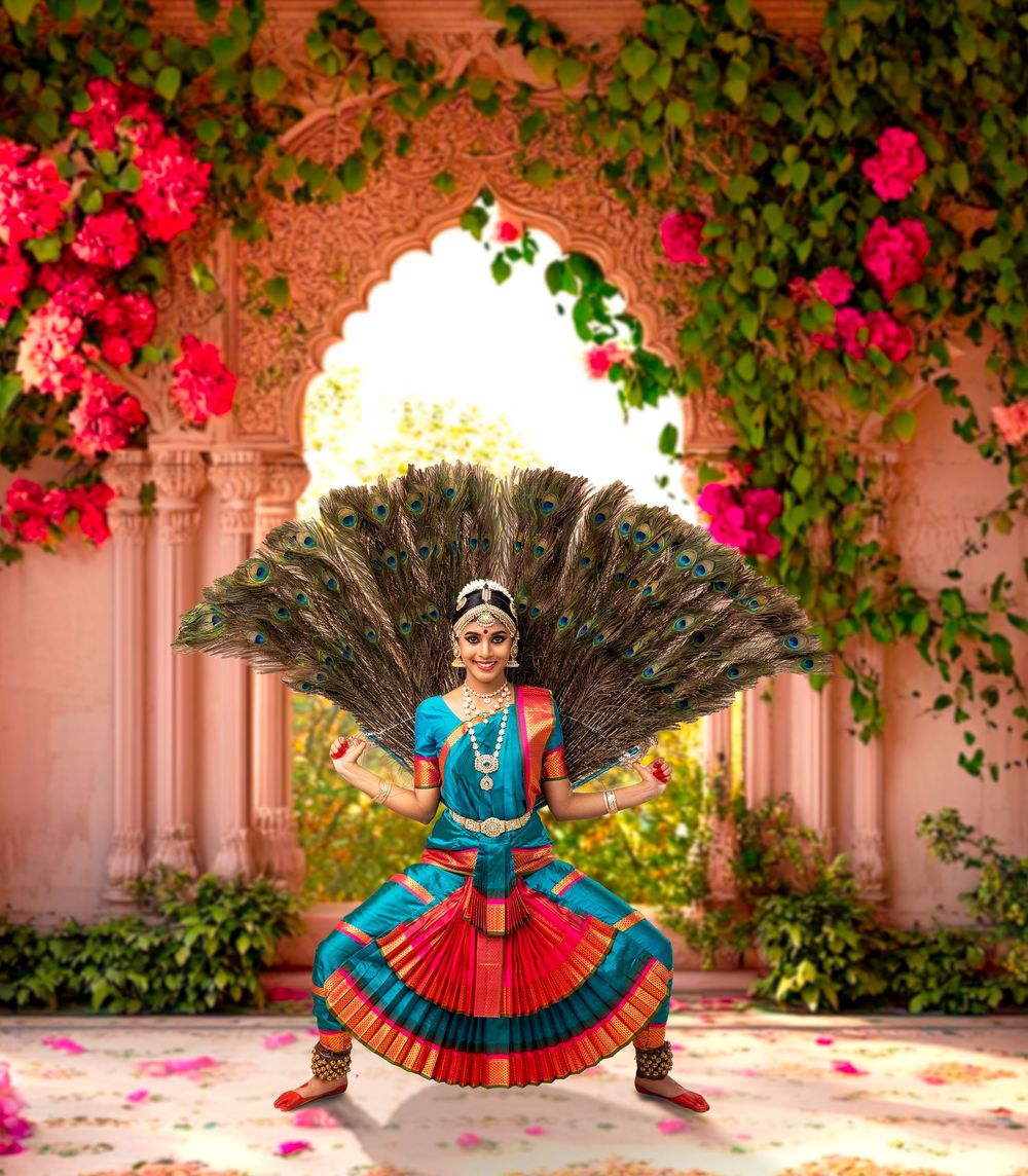 Classical Indian dancer in a vibrant blue and red costume performs with a peacock feather fan against a floral archway.