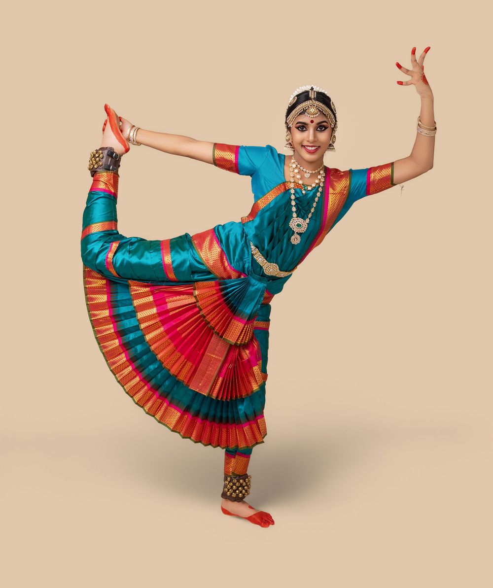 Dancer performs a traditional Bharatanatyam dance pose in turquoise and red costume against a beige backdrop.