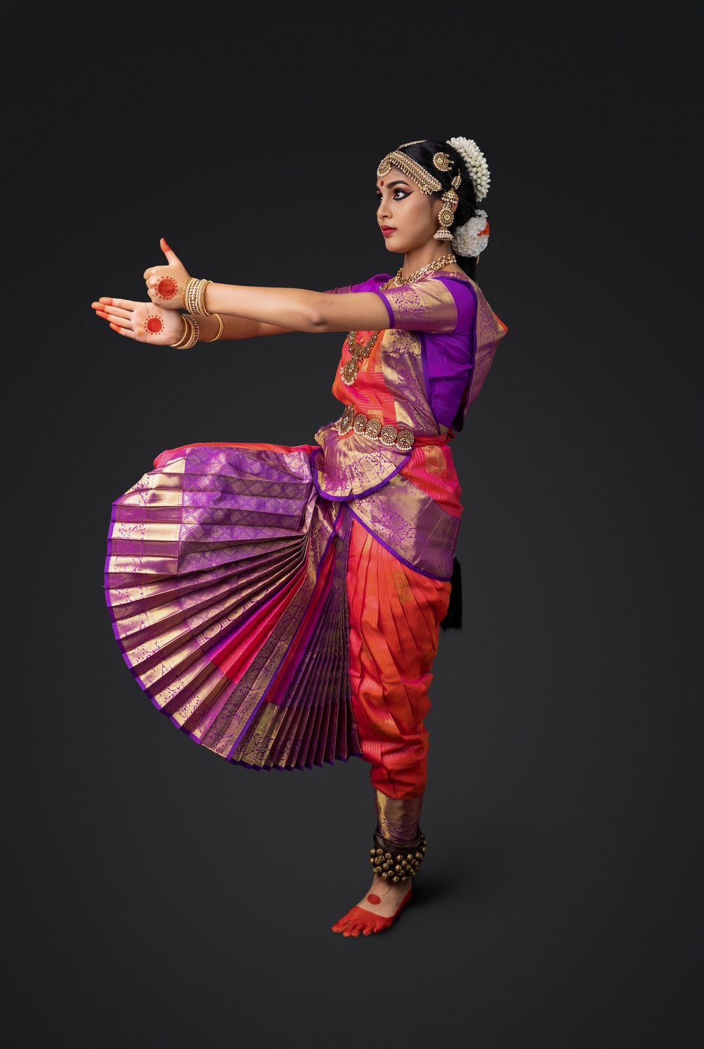 Classical Indian dancer in vibrant purple and orange sari performs traditional Bharatanatyam pose against dark background.