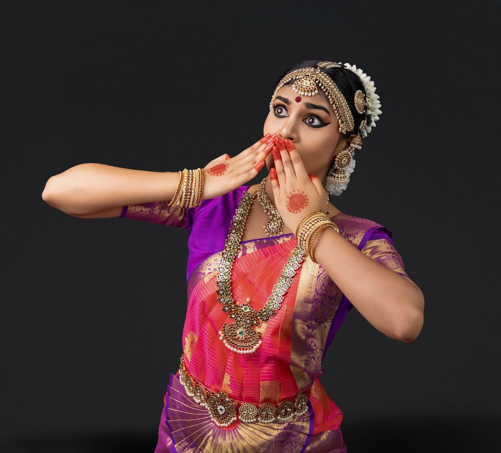 Classical Indian dancer in traditional pink and purple attire performs expressive hand gestures against dark background.