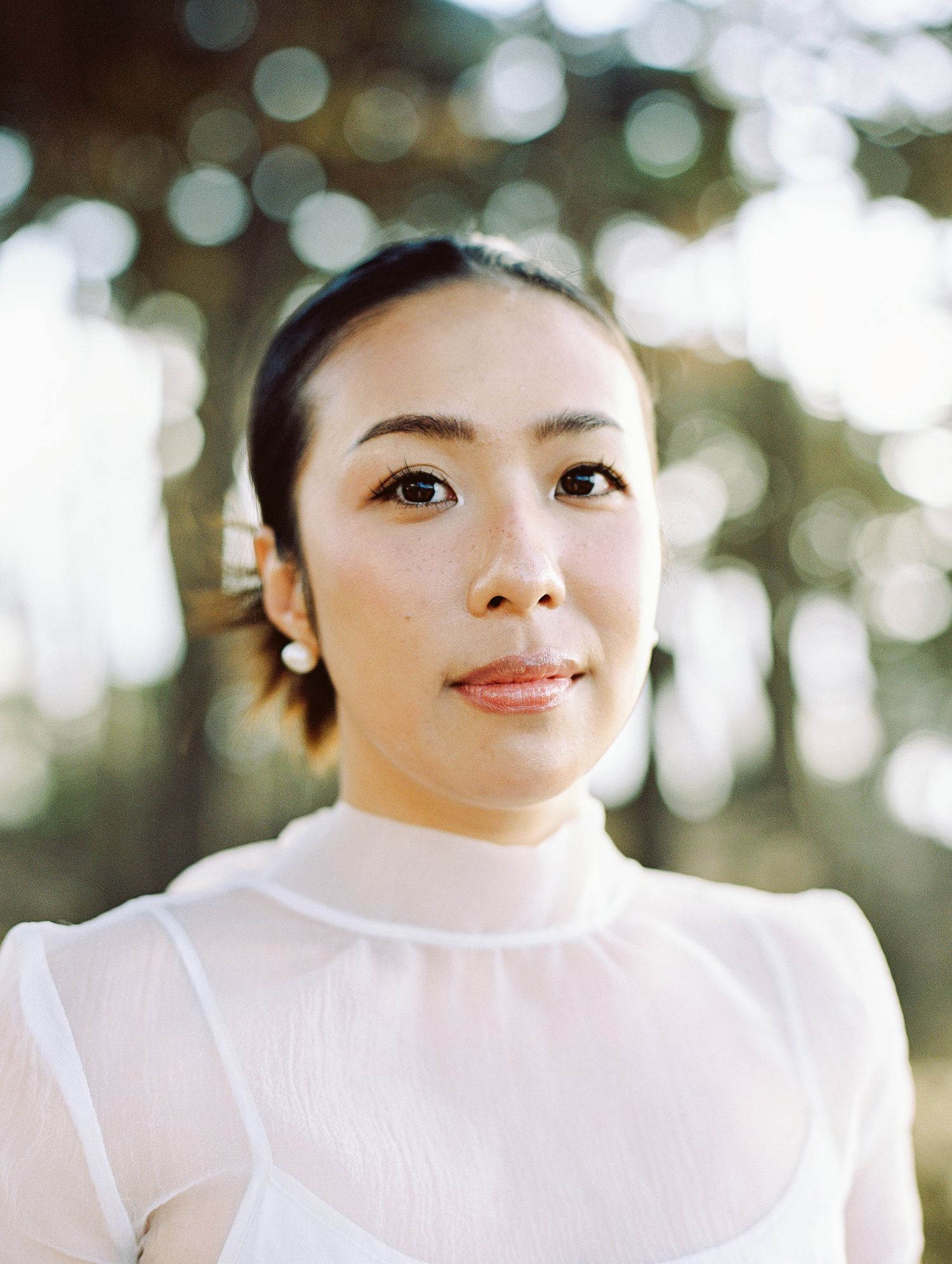 Portrait of someone in a white high-neck blouse with delicate earrings against a soft bokeh background.