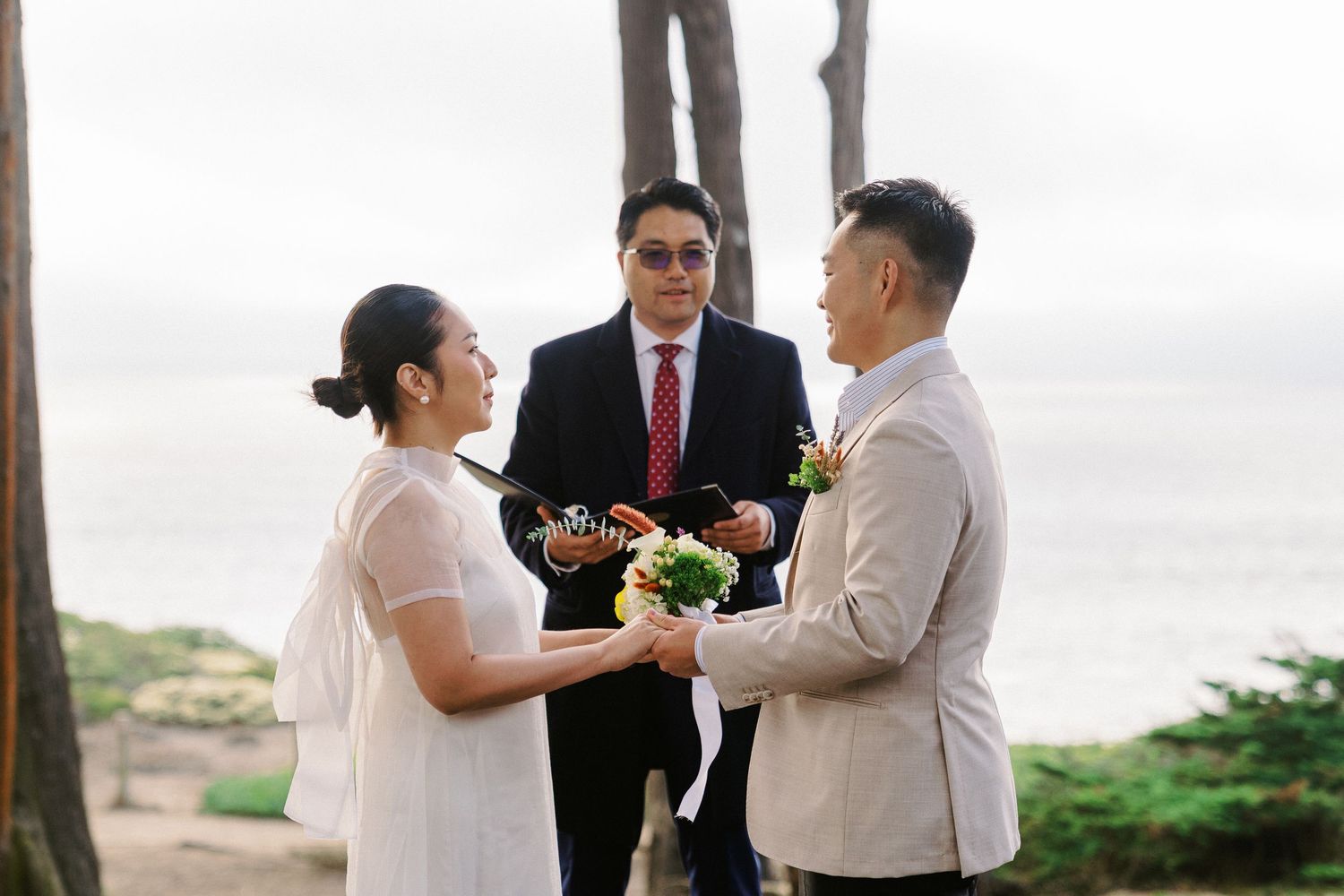 Wedding ceremony taking place outdoors on a beach with palm trees and ocean views.