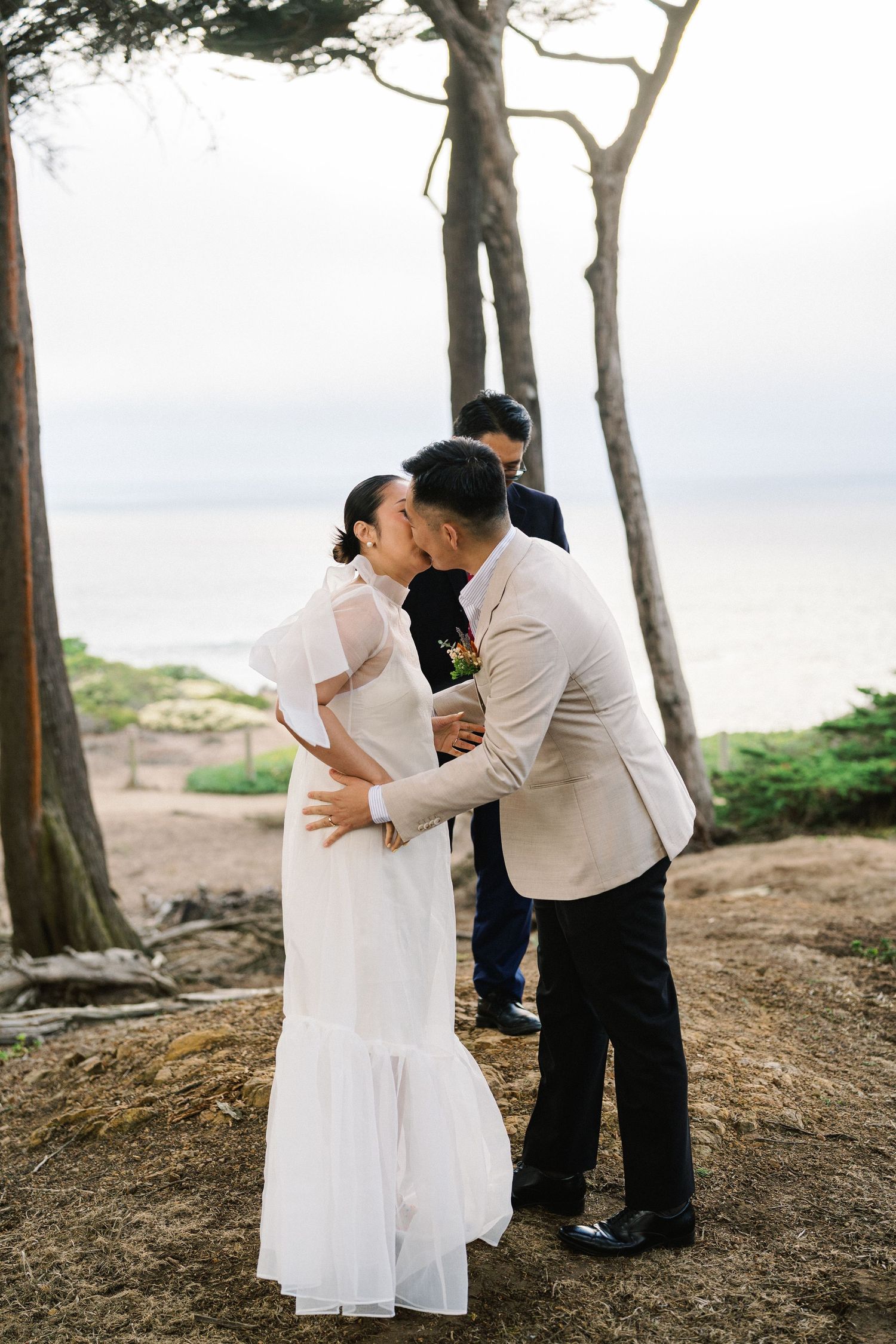 A couple in white and black attire embrace in a romantic photoshoot among tall trees by the coastline.